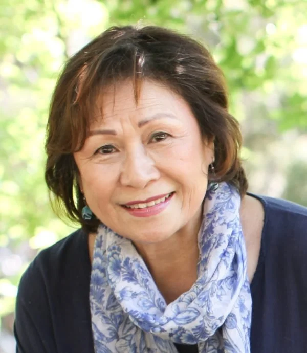 A smiling middle-aged woman with short brown hair, wearing a blue top and a blue and white patterned scarf, outdoors with a background of green foliage.