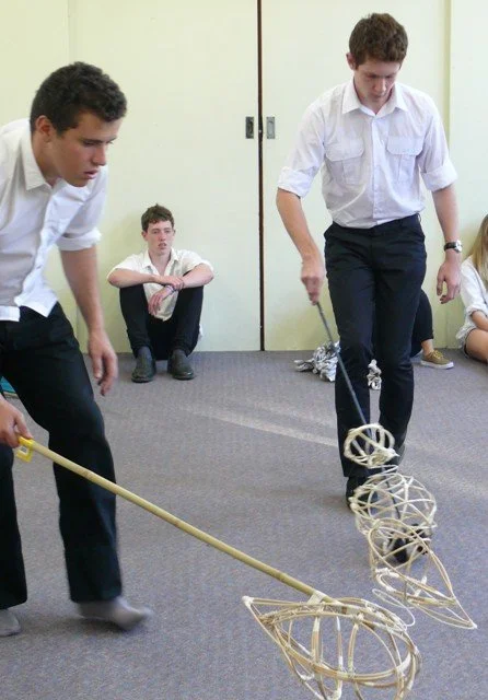 Two young men in white shirts and dark pants play hockey with wooden sticks and a ball in a room with yellow walls. Two children sit on the floor in the background, observing.
