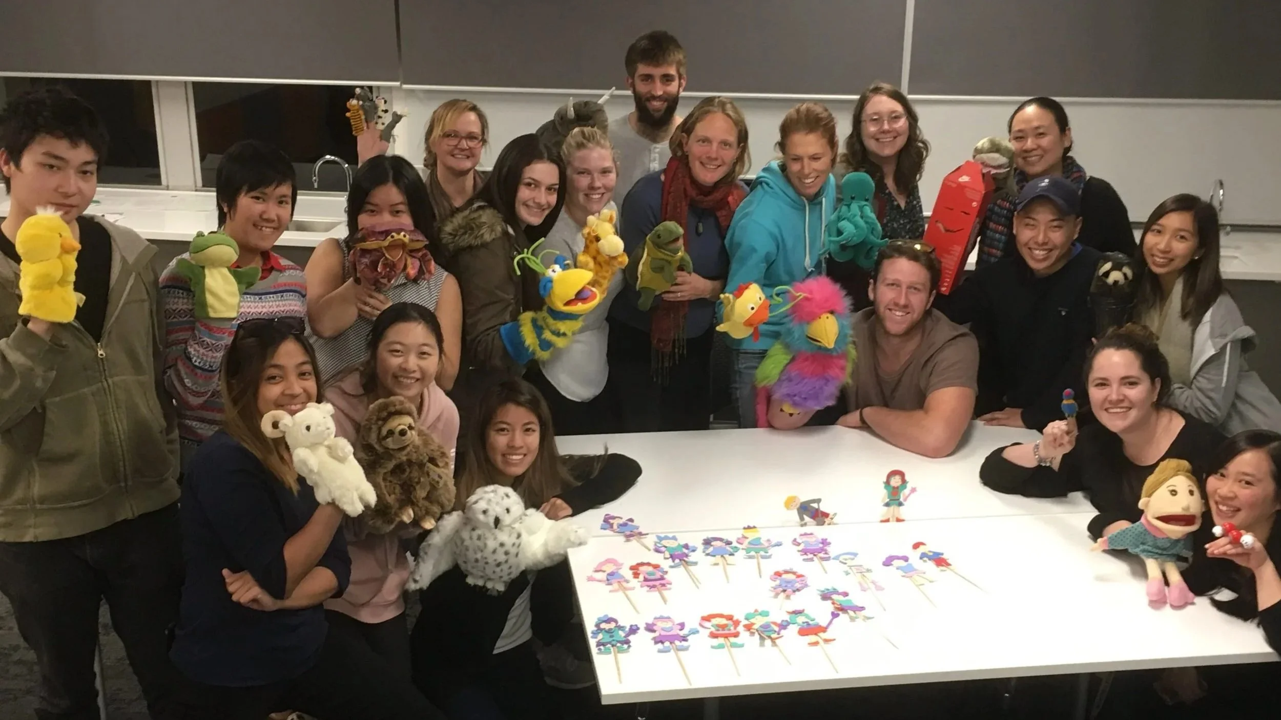 Group of people in a classroom holding puppets and puppetry supplies, with puppets on the table.