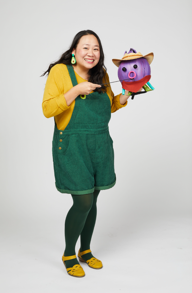 A woman dressed in a yellow long-sleeve top, green overalls, green tights, and yellow shoes, smiling while holding a colorful paper craft of a pig wearing a hat and scarf.