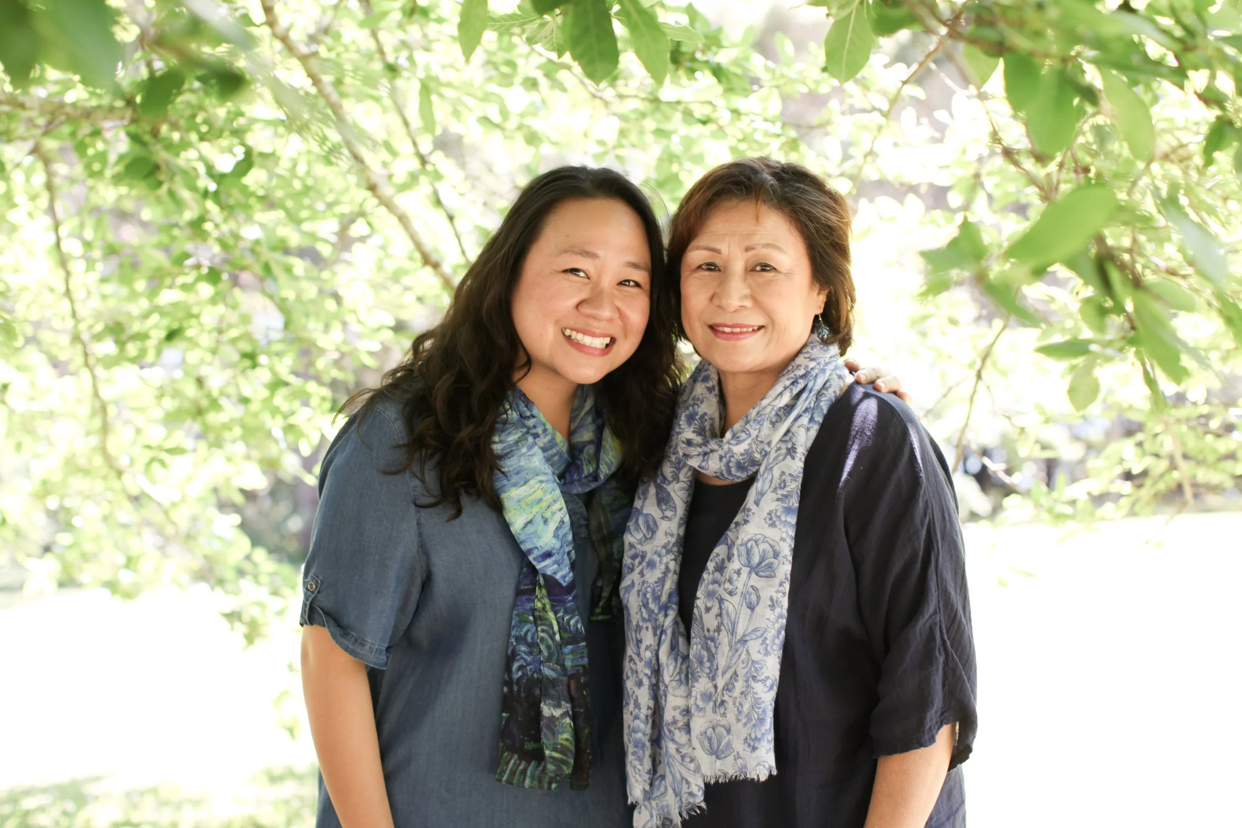 Two women smiling and hugging outdoors under green leafy trees.