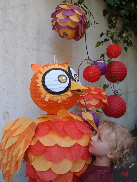 A child with blonde hair and a maroon shirt touching a colorful owl-shaped lantern decoration made of paper, with hanging lanterns and decorative elements in the background.