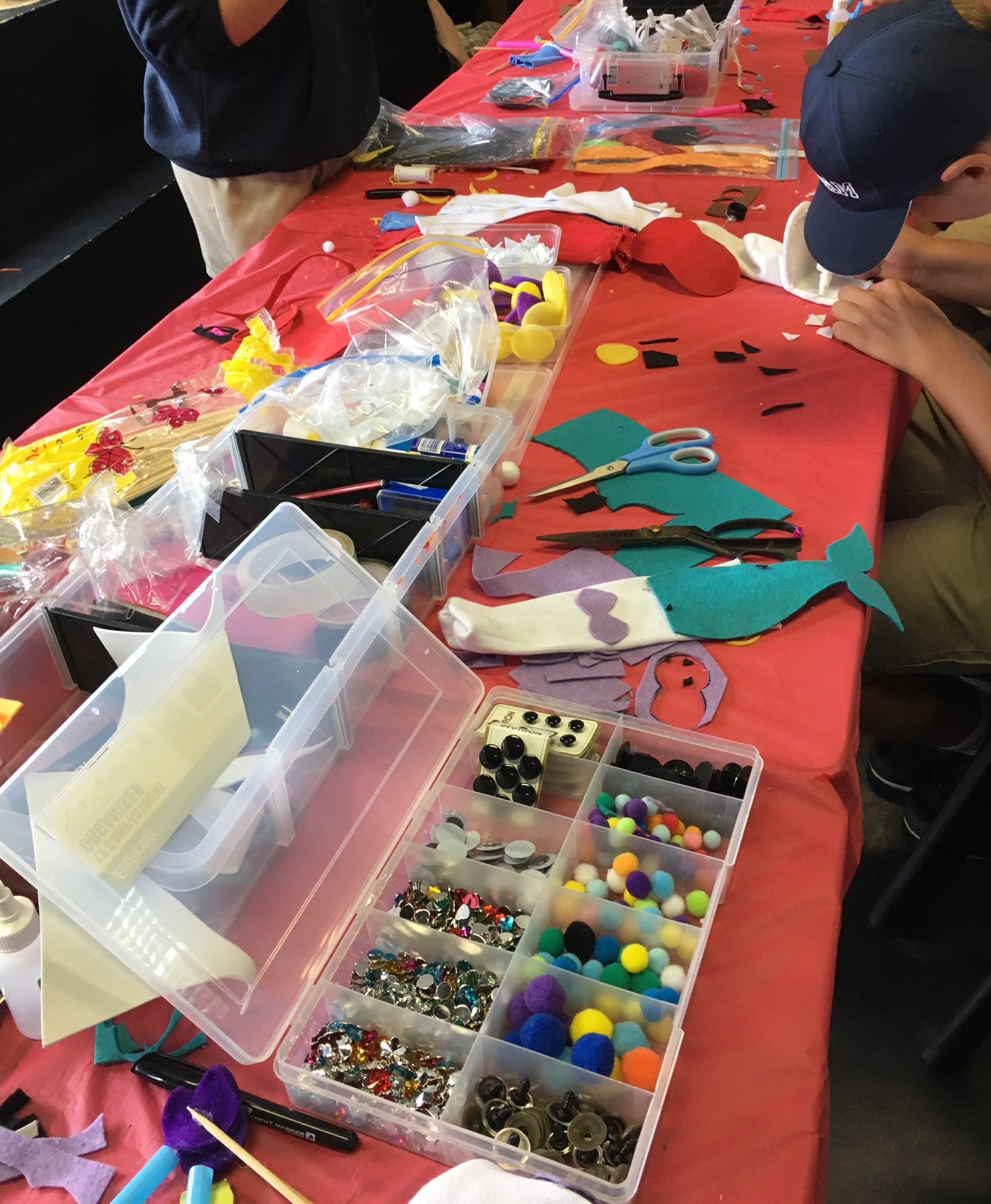 A crafting table covered with a red tablecloth, filled with various supplies like buttons, pom-poms, felt, scissors, markers, and decorative items for arts and crafts.