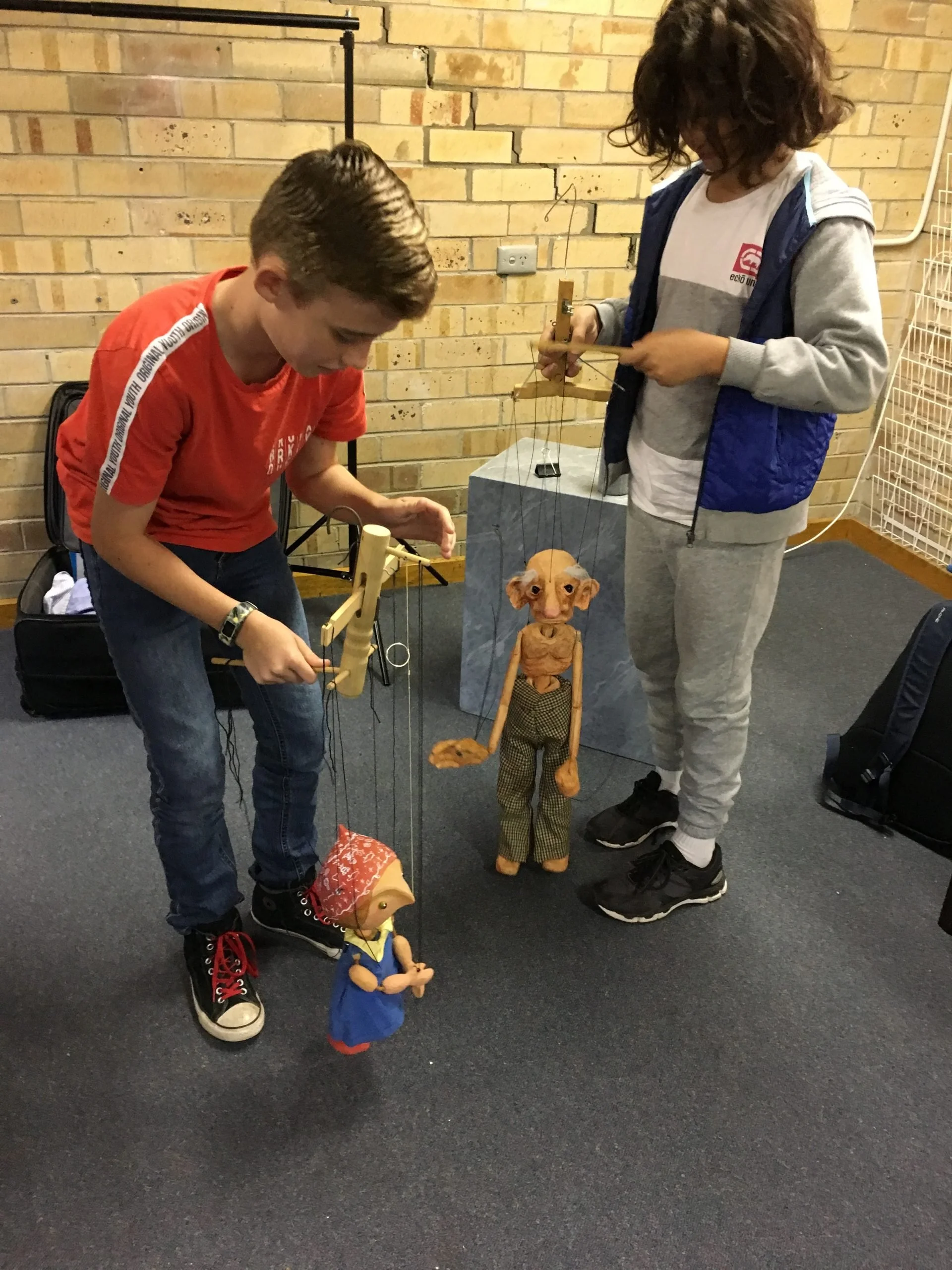 Two children playing with marionette puppets indoors, with a brick wall in the background. One child is wearing a red t-shirt and jeans, the other a gray sweatshirt with a blue vest and gray sweatpants.