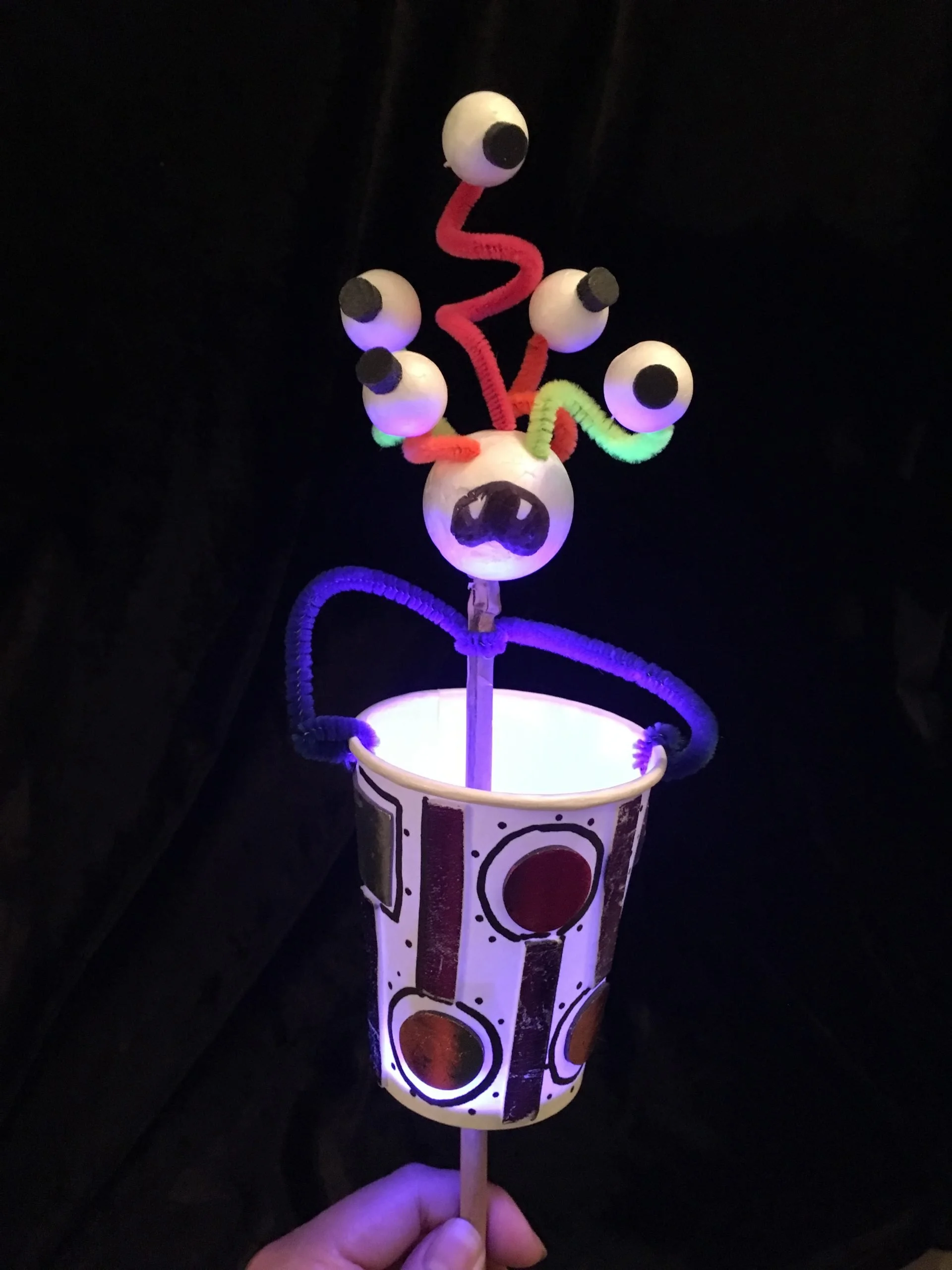 Hand holding a light-up puppet resembling a cartoonish alien with six eyeballs on flexible stalks and a white body with black circles, set against a dark background.