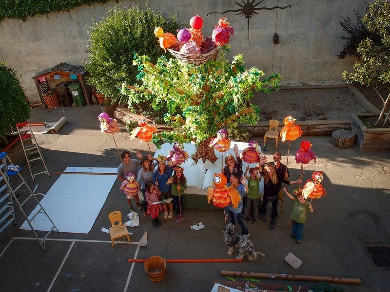 Group of people and a dog arranged around a large decorative tree with colorful owl and bird ornaments in an outdoor yard.