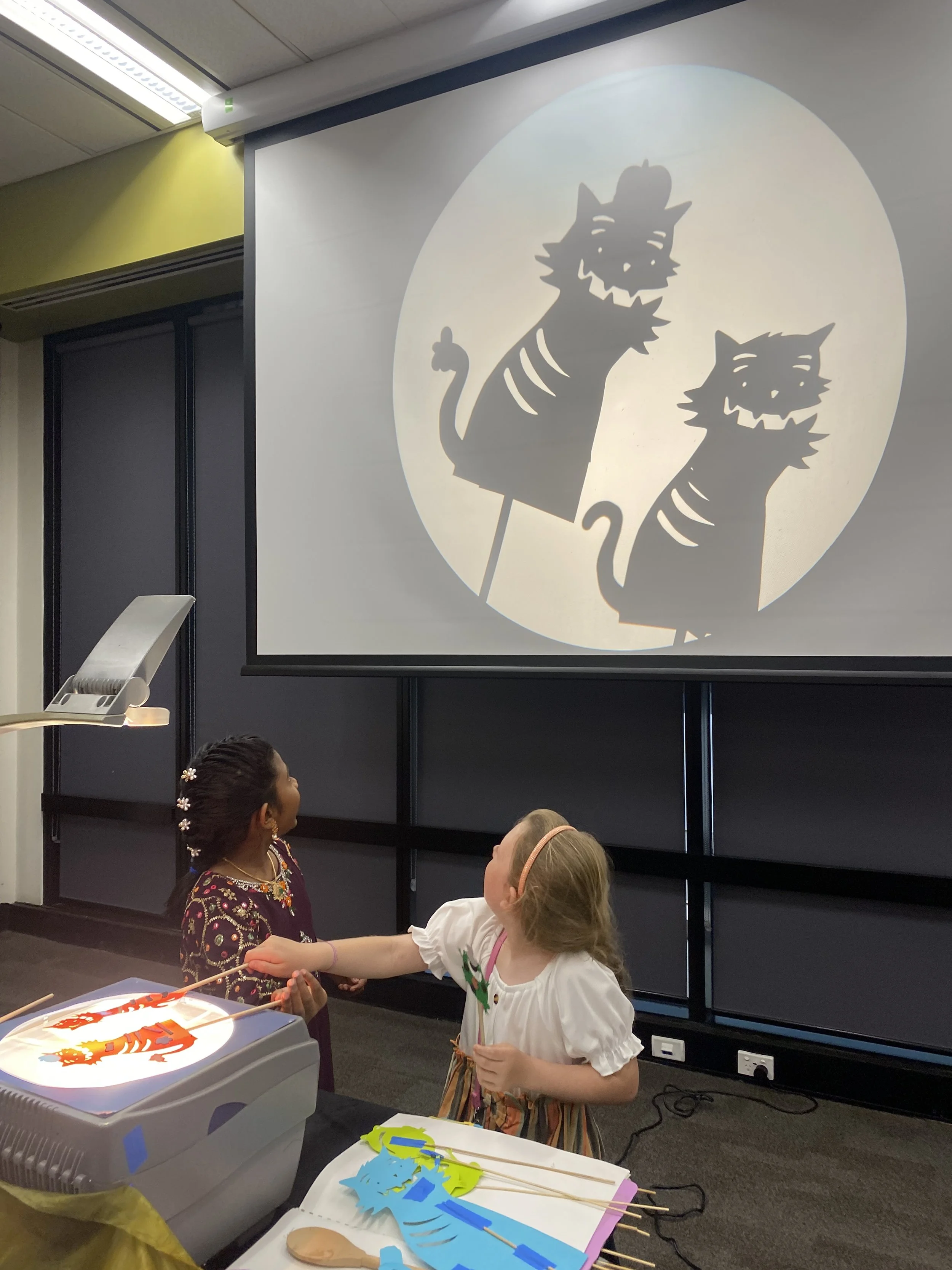 Two children are engaged in a craft activity, with one girl holding a paper cut-out and the other girl reaching towards it, in a room with a large screen projecting silhouettes of a cat and a mouse.