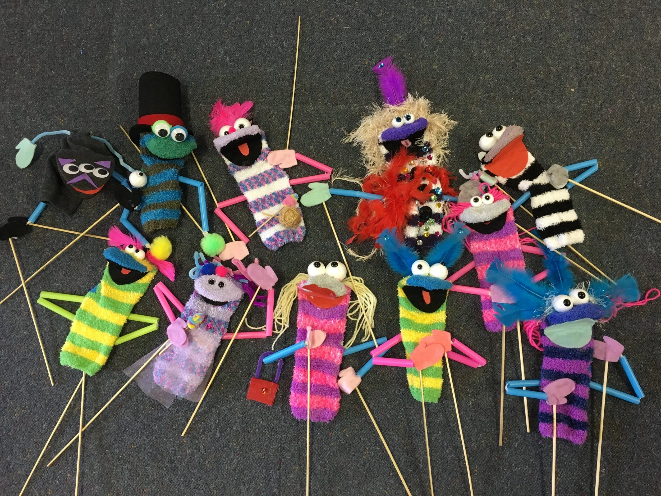 Collection of colorful sock puppets with googly eyes, feathers, pipe cleaners, and pom-poms arranged on a dark carpeted surface.