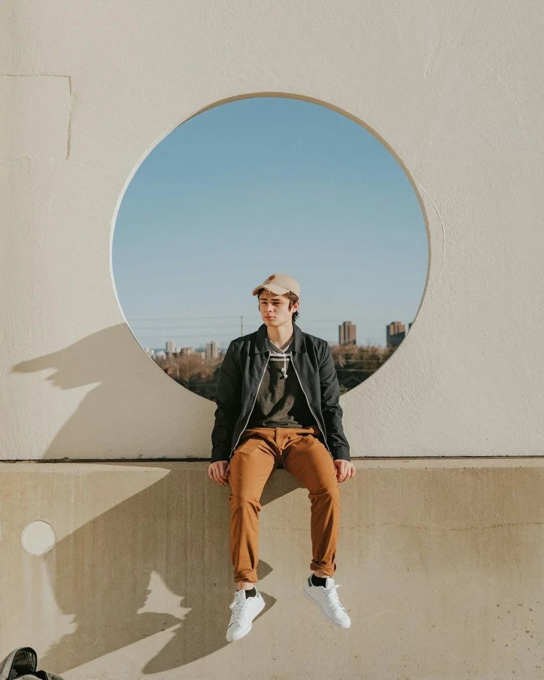 A young man sitting on a concrete ledge in front of a circular window in a white wall, with a city skyline and power lines visible in the background, on a sunny day.
