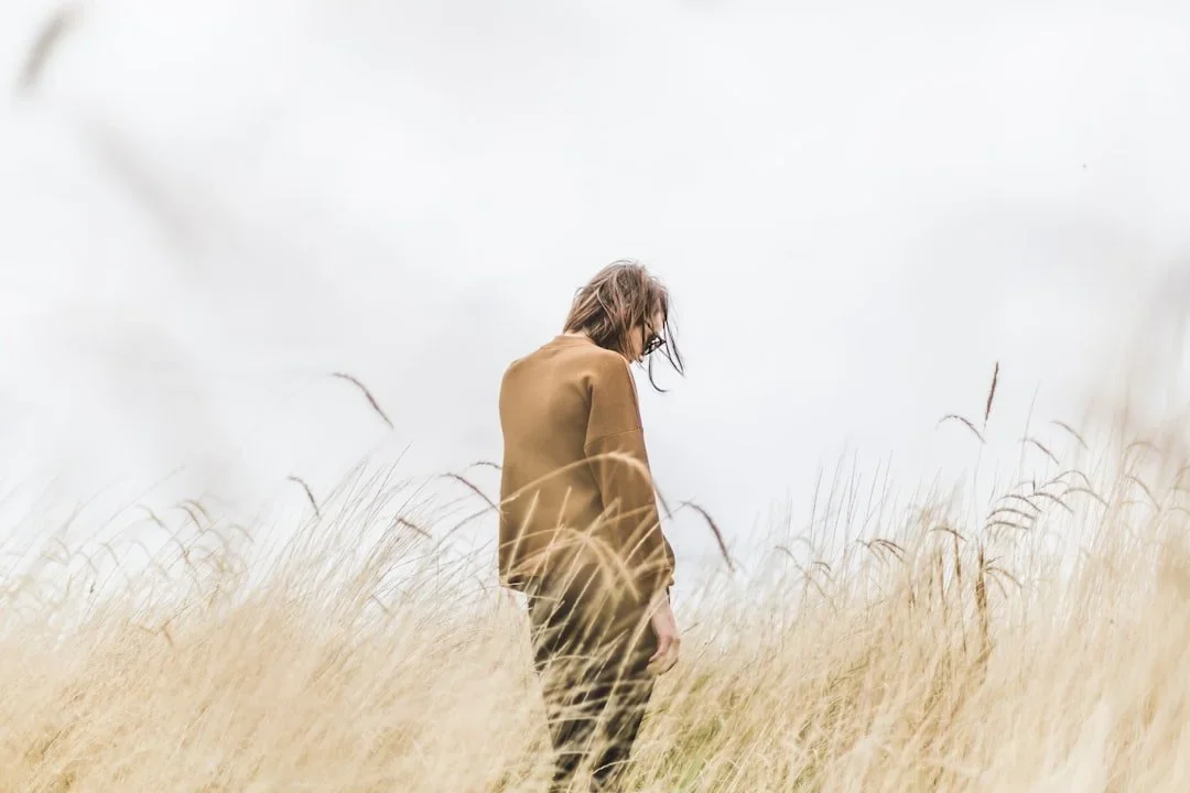 A woman with shoulder-length hair, wearing a tan jacket, standing in a field of tall, golden grass, looking down with her head lowered.