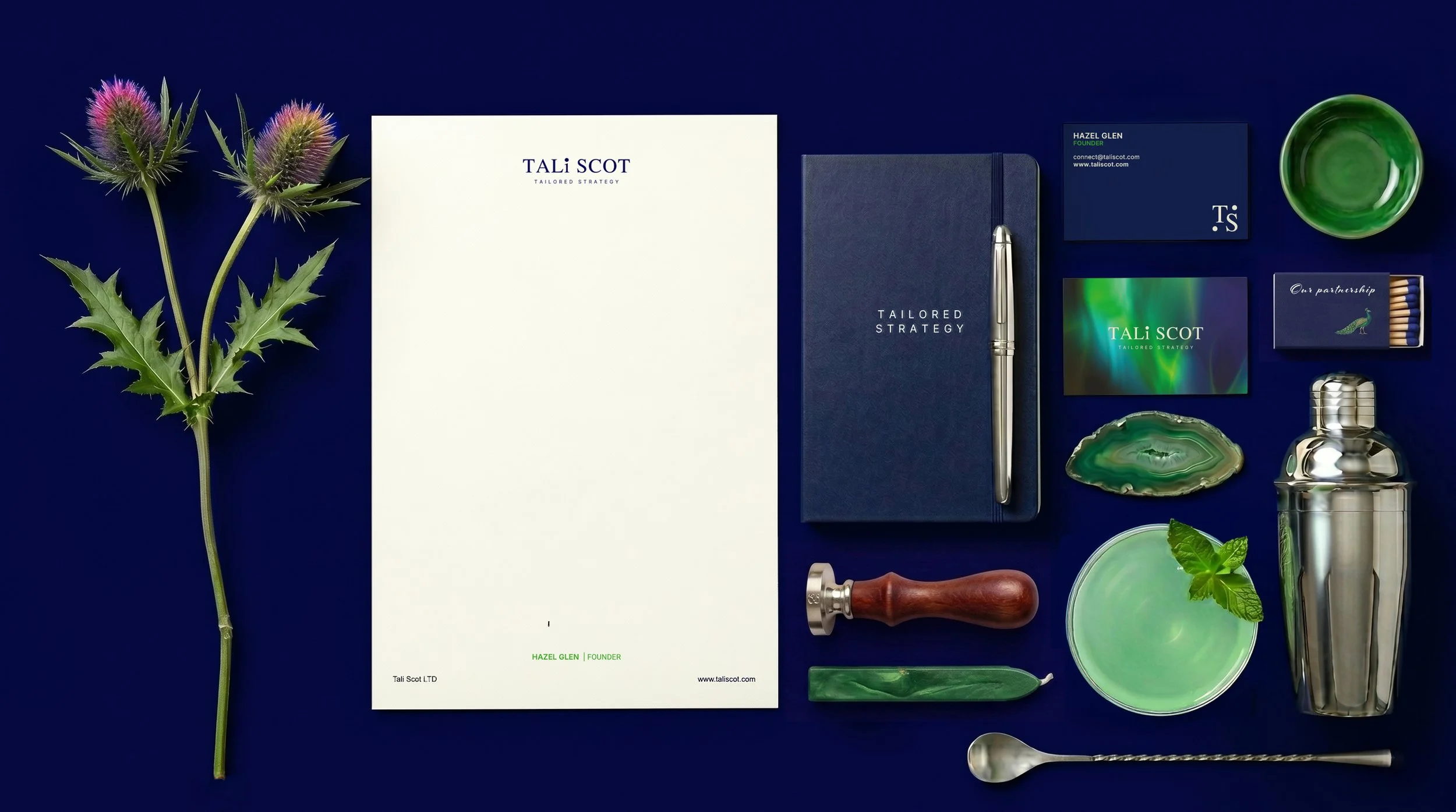A flat lay of Tali Scot branded stationery, business cards, a cocktail shaker, a green cocktail, and Scottish thistles arranged on a dark blue background.