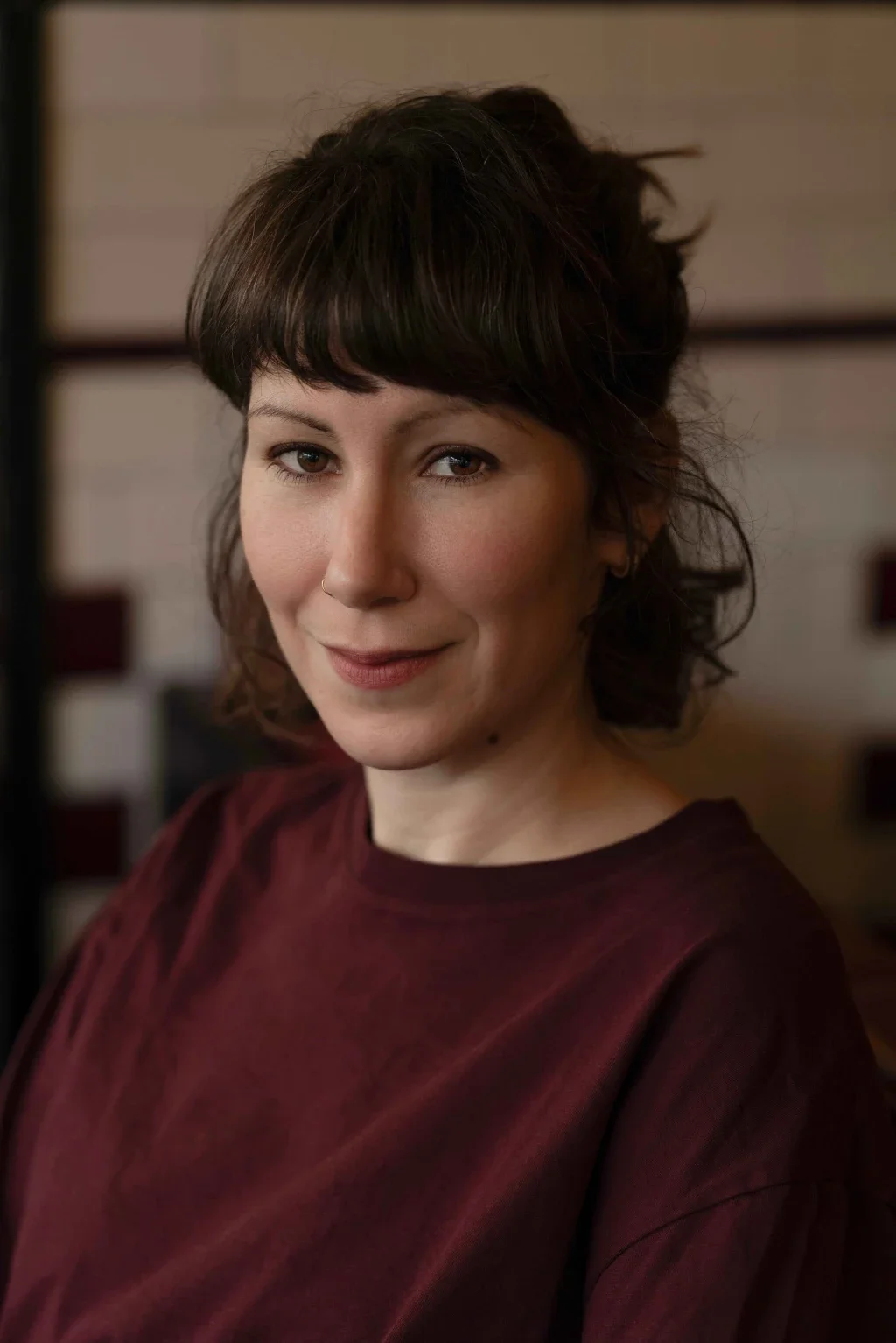 Portrait d'une femme avec des cheveux bruns bouclés, portant un haut bordeaux, souriant légèrement, en intérieur.