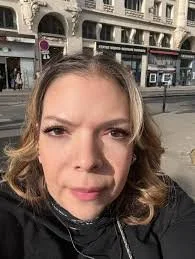 Une femme souriante avec des cheveux bruns, prenant un selfie dans une ville urbaine.