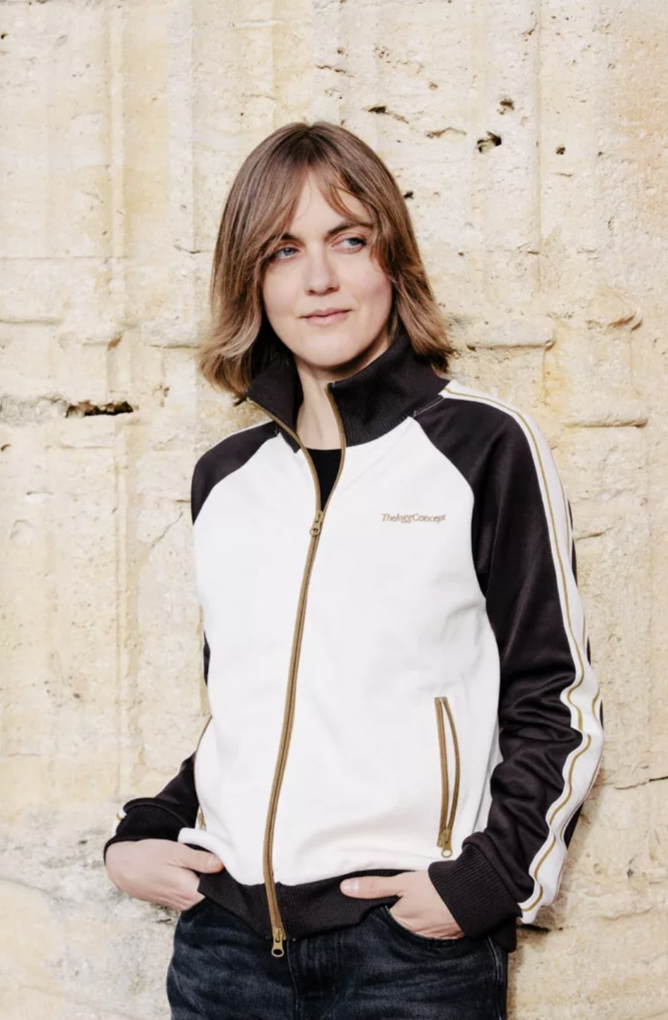 Une jeune femme portant une veste de sport noire et blanche avec des détails dorés, posant devant un mur en pierre beige.