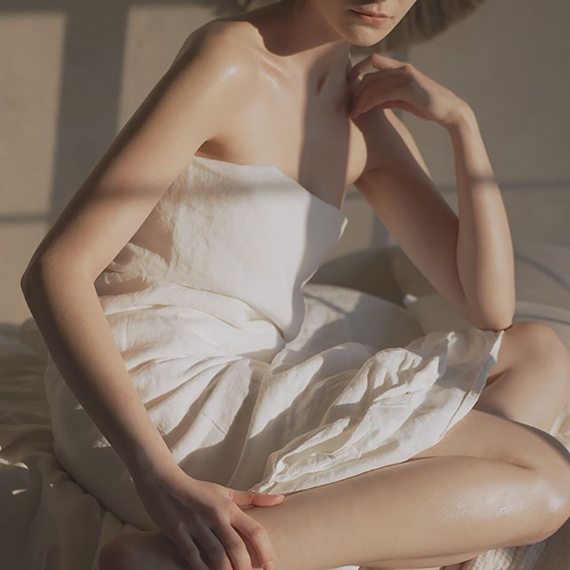 A woman sitting on a bed, wrapped in a white towel, with sunlight casting shadows on her bare shoulder and arm.
