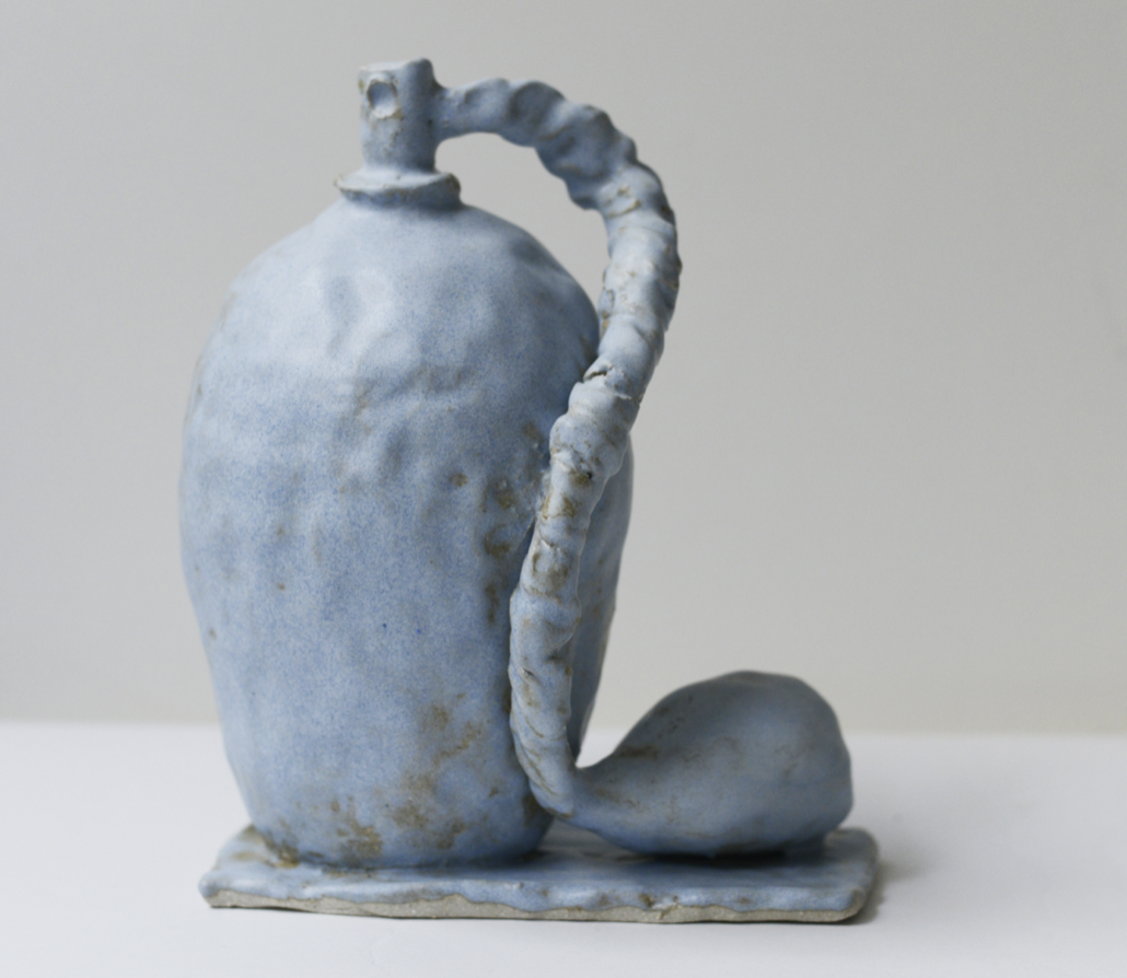 Une sculpture en céramique représentant une bouteille bleue avec un bouchon, accompagnée d'une cuillère ou d'une spatule, sur une surface plate.