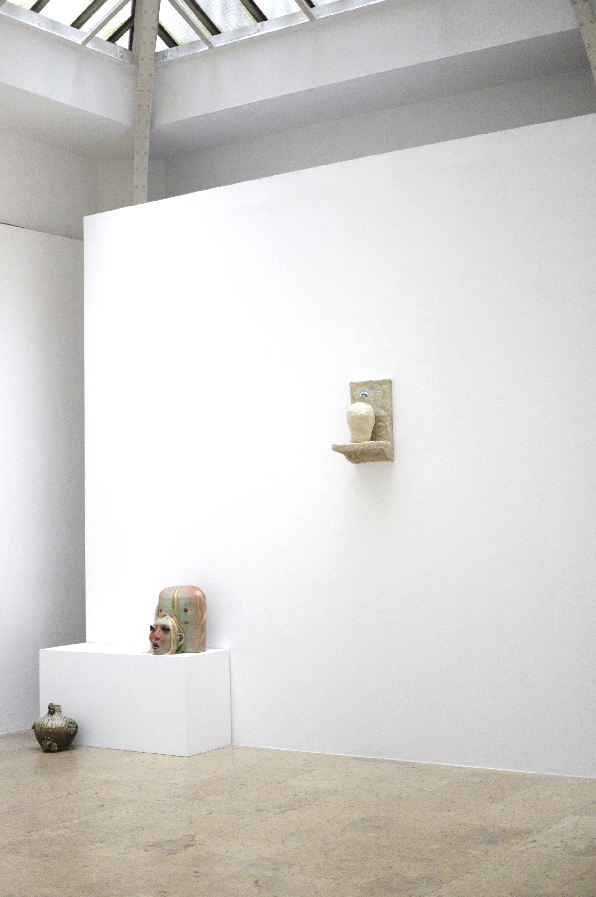 Exposition d'art contemporain avec des sculptures et un masque de visage féminin sur un socle, dans une salle à blanc avec un mur blanc et un plafond en verre.
