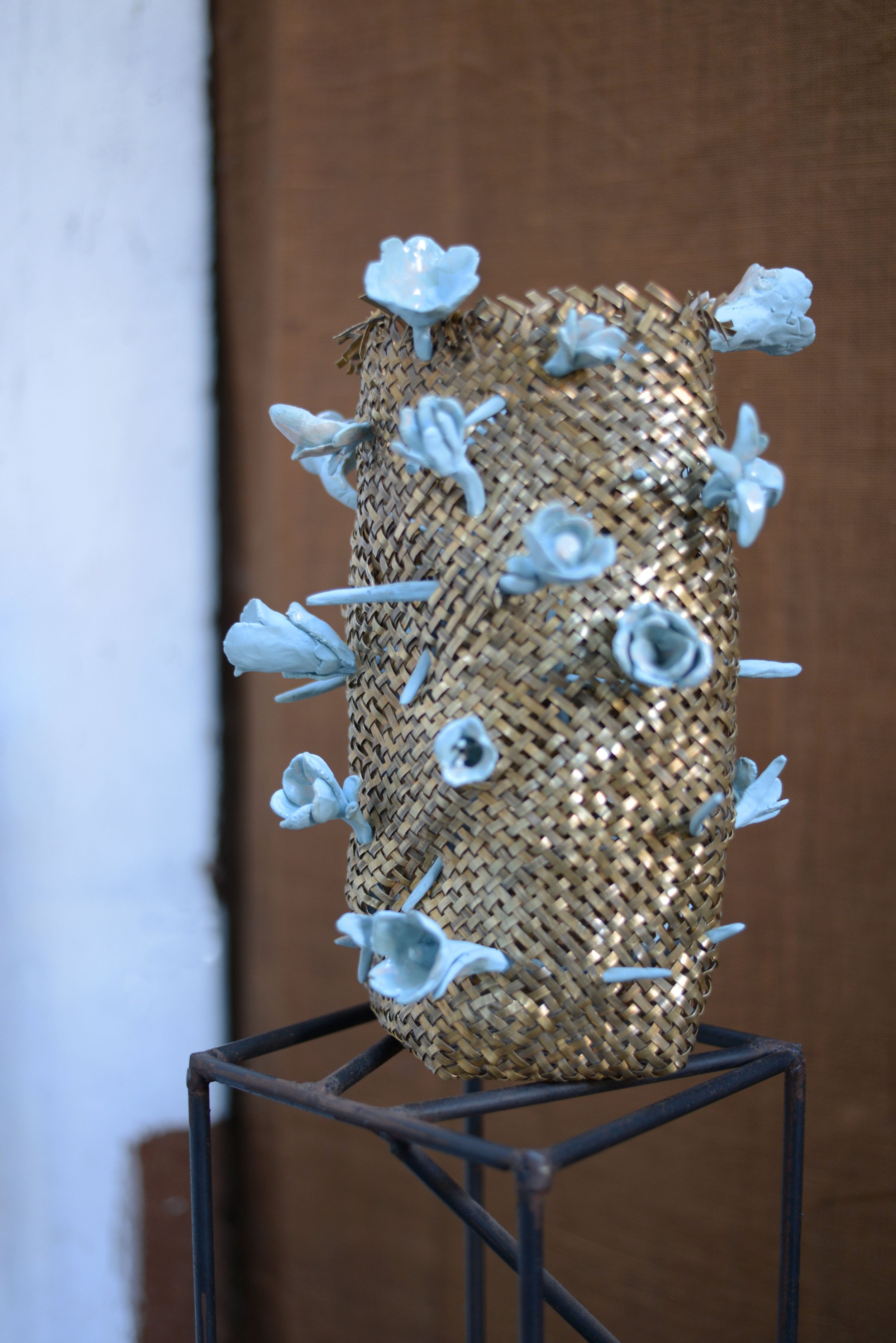 Une sculpture composée d'un panier en osier contenant des fleurs en céramique blanche, placée sur une structure métallique.