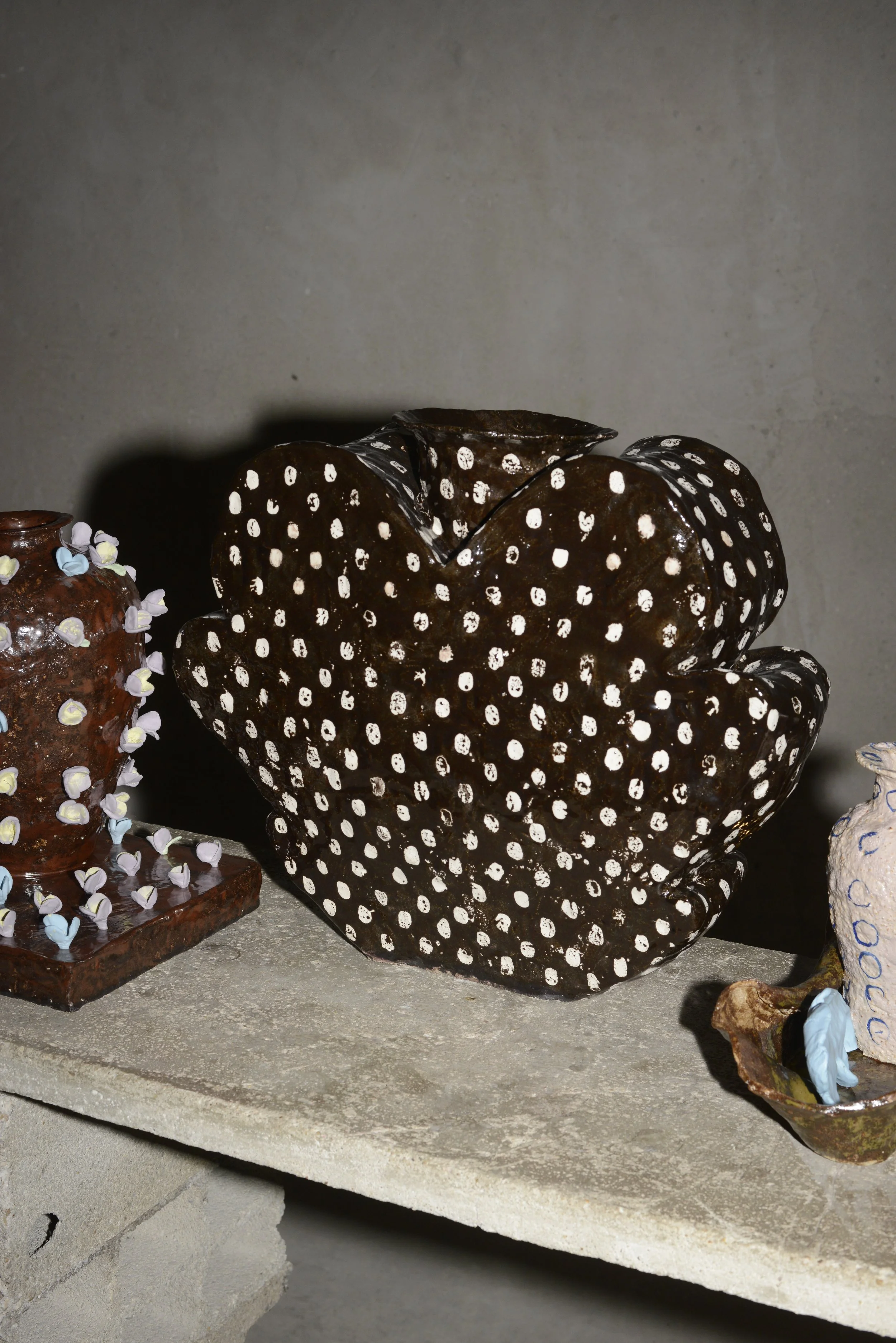 Vase en céramique noire avec motifs de points blancs, autre poterie à gauche décorée de petites fleurs colorées, et autres objets en céramique à droite. Plats posés sur une étagère en pierre ou béton.