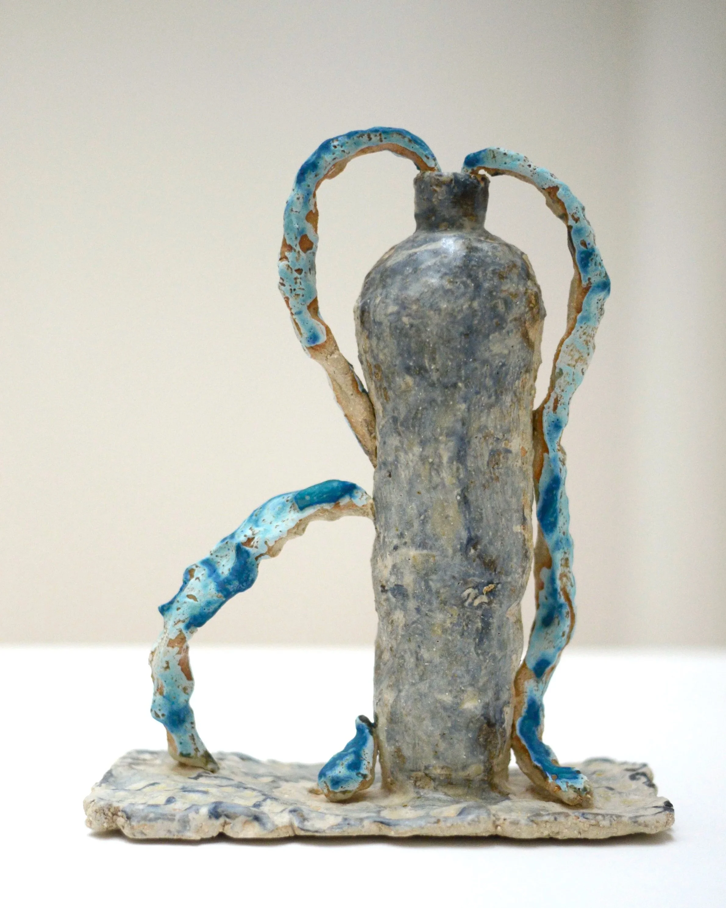 Une sculpture artistique de forme humanoïde avec des éléments en terre cuite, comportant des détails en bleu et beige, reposant sur une base en terre.