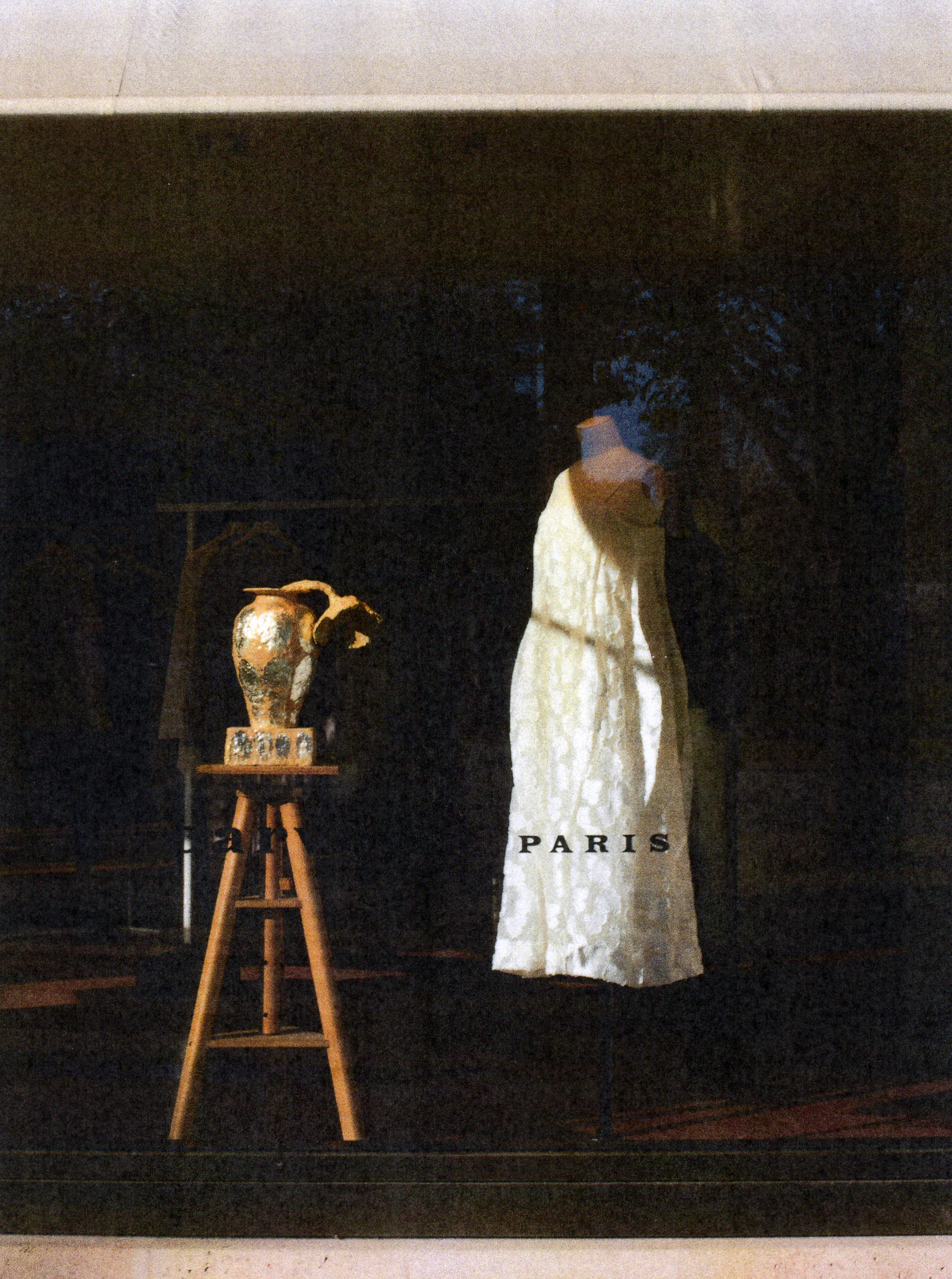 Intérieur sombre avec une toile blanche portant le texte 'PARIS'. Sur la gauche, un chevalet avec une vase ancienne en céramique. En arrière-plan, un tableau ou un décor sombre avec des feuilles d'arbres visibles.