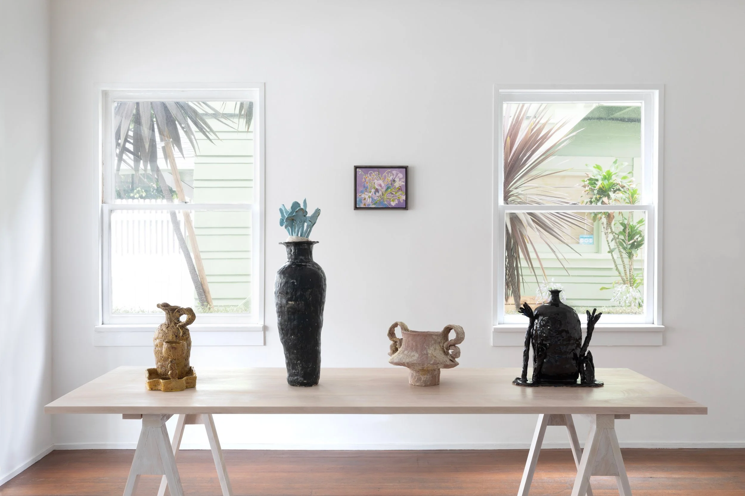Une table en bois clair avec plusieurs sculptures en céramique, dont une noire et une beige, devant deux fenêtres laissant entrer de la lumière, dans une pièce épurée.