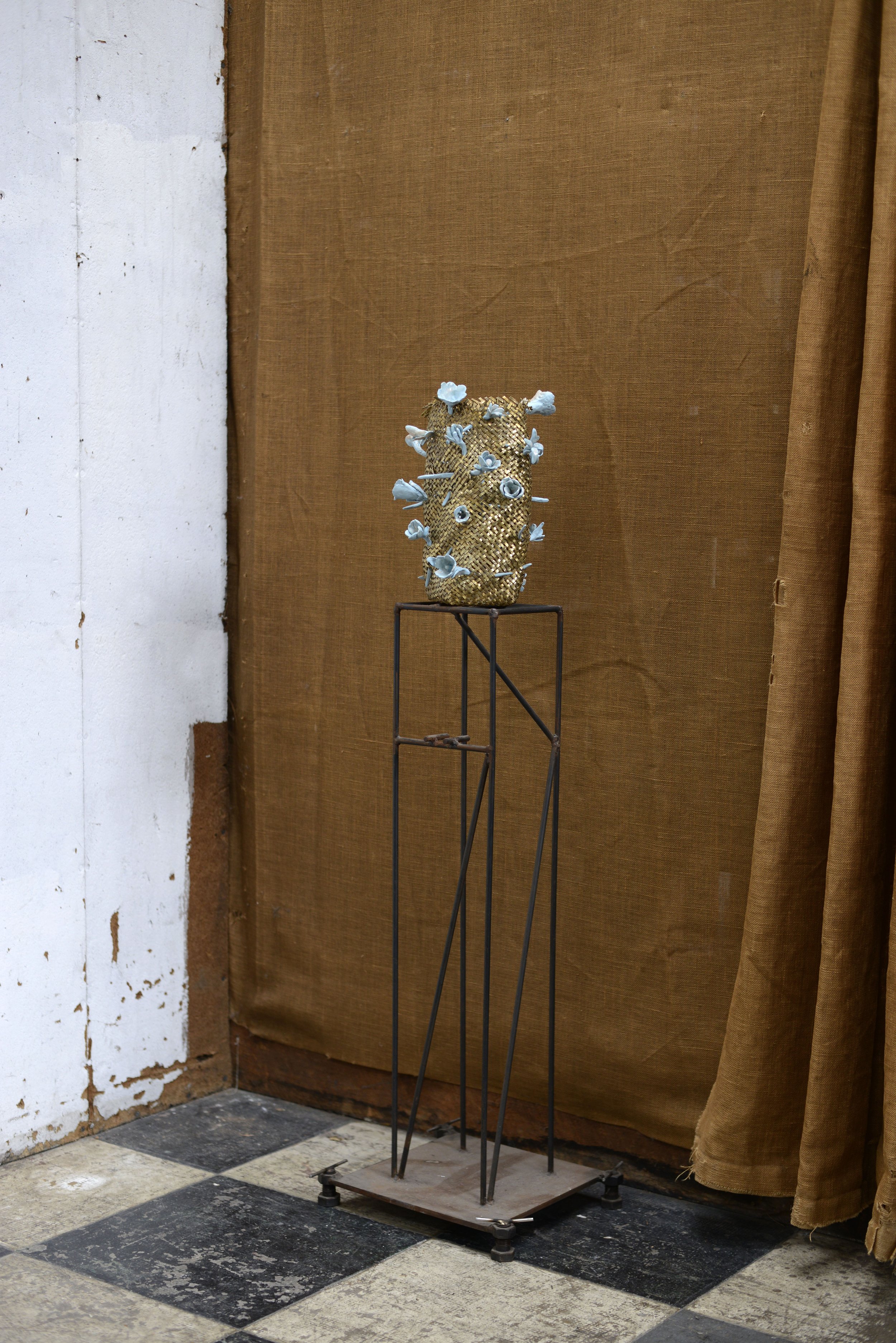 Vase or sculpture en forme de pot avec décoration de fleurs blanches, placé sur un support en métal dans un espace intérieur avec un fond en tissu marron et un mur en bois, sol en carreaux noir et blanc.