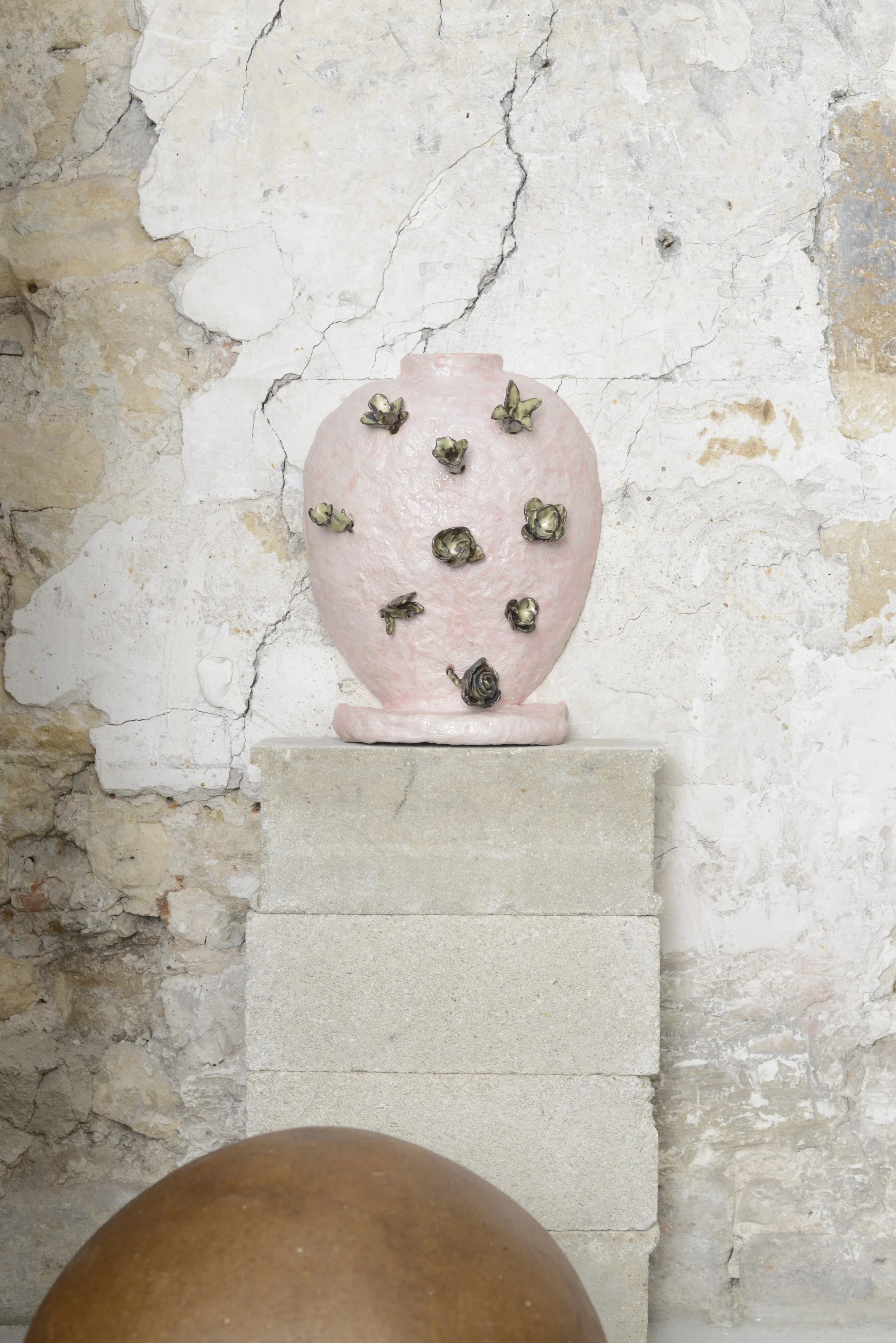 Une installation artistique représentant un vase en pierre rose avec des fleurs métalliques noires, posé sur un socle en pierre, contre un mur en pierre aux coloris neutres et diverses fissures.