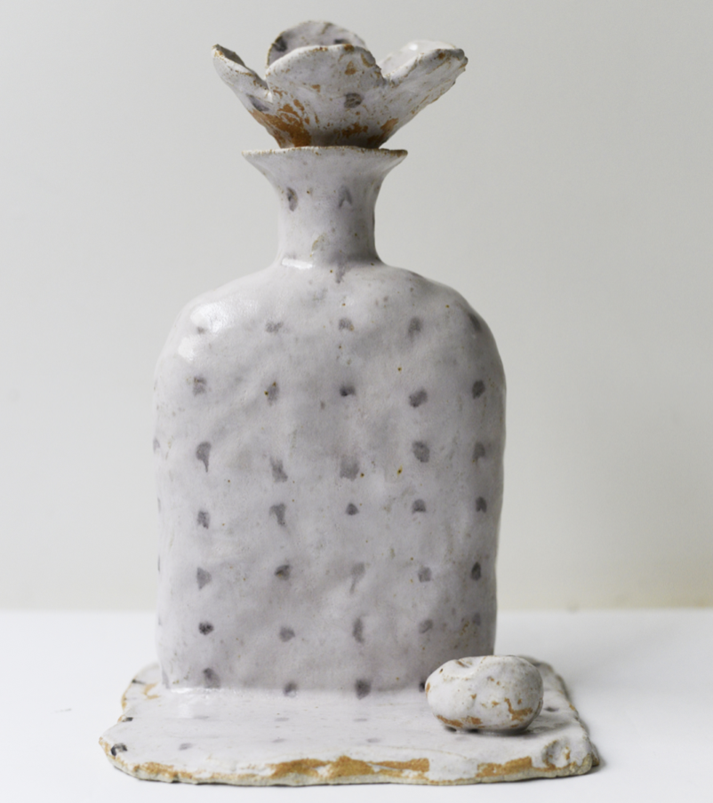 Une sculpture en céramique représentant un flacon avec une forme de fruit ou de fleur en haut, de couleur blanc avec des taches brunes, posée sur une surface plate également blanche avec des taches brunes.