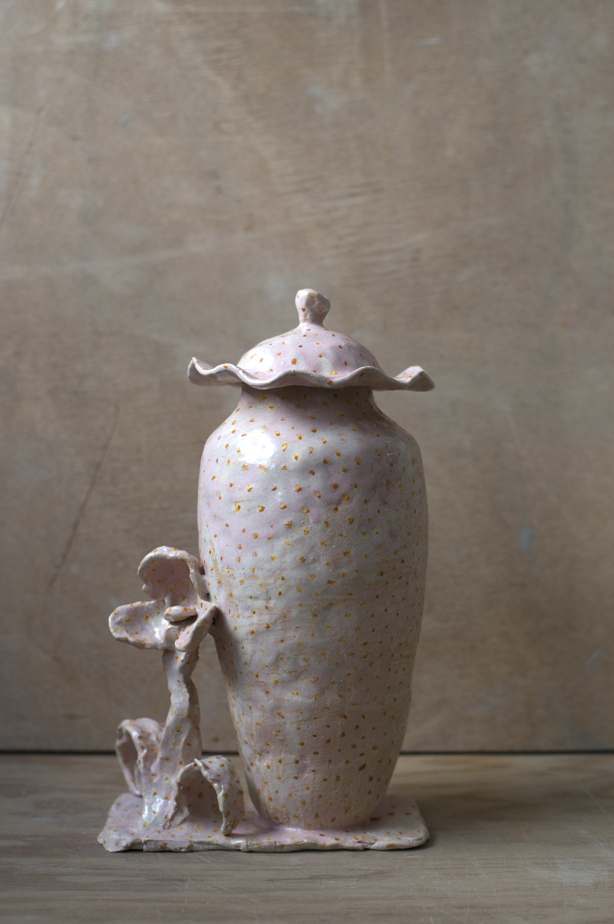 Une sculpture en céramique de forme vases avec des détails en forme de feuilles et de fleurs, ornée de motifs pointillés roses et dorés sur une surface blanche, posée sur une table en bois claire.