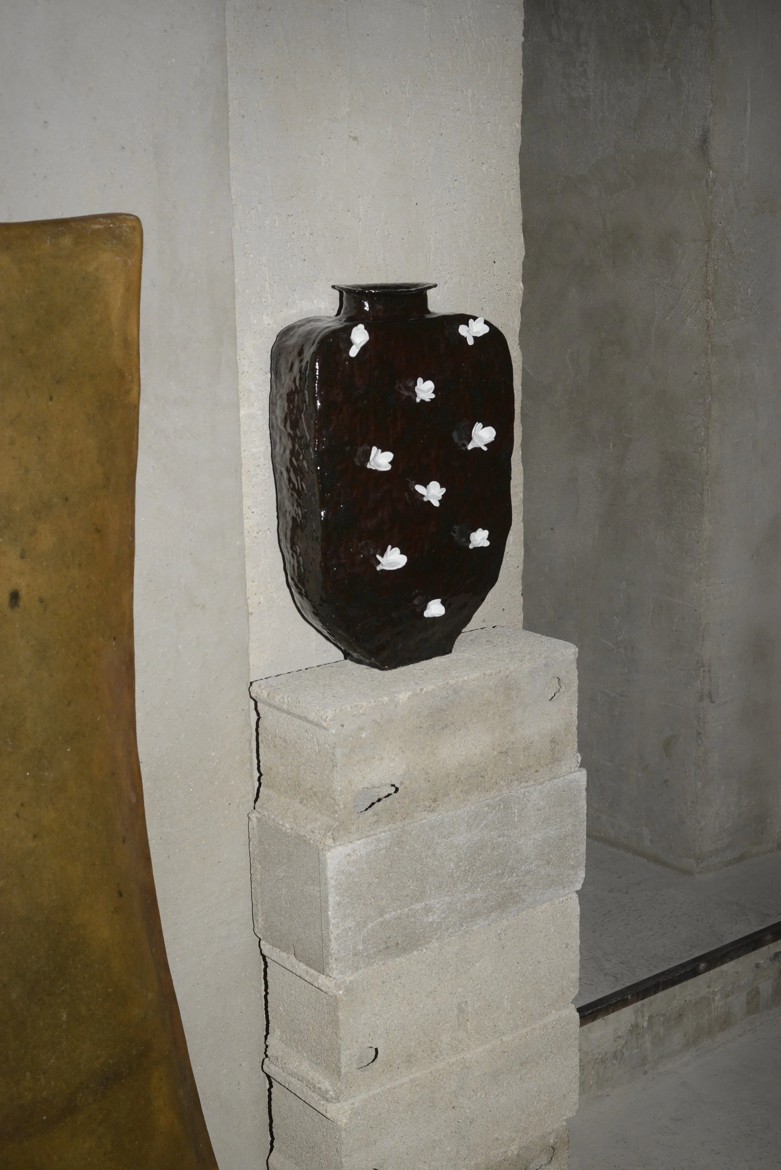 Une poterie noire décorée de petites fleurs blanches, placée sur une étagère en pierre, avec un fond de murs en béton et une autre pièce de poterie beige partiellement visible à gauche.