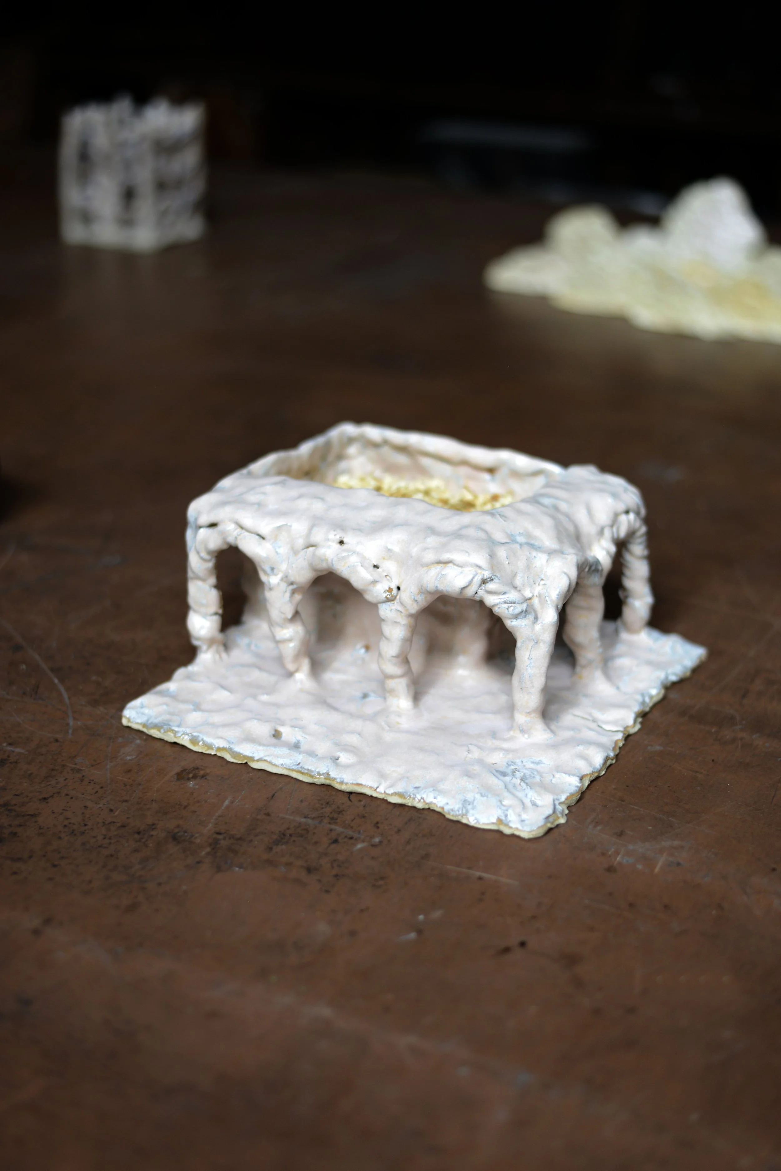 Une sculpture en forme de bâtiment avec une structure complexe en matériaux blanc, placée sur une surface en bois. En arrière-plan, il y a d'autres formes semblables, mais floues.
