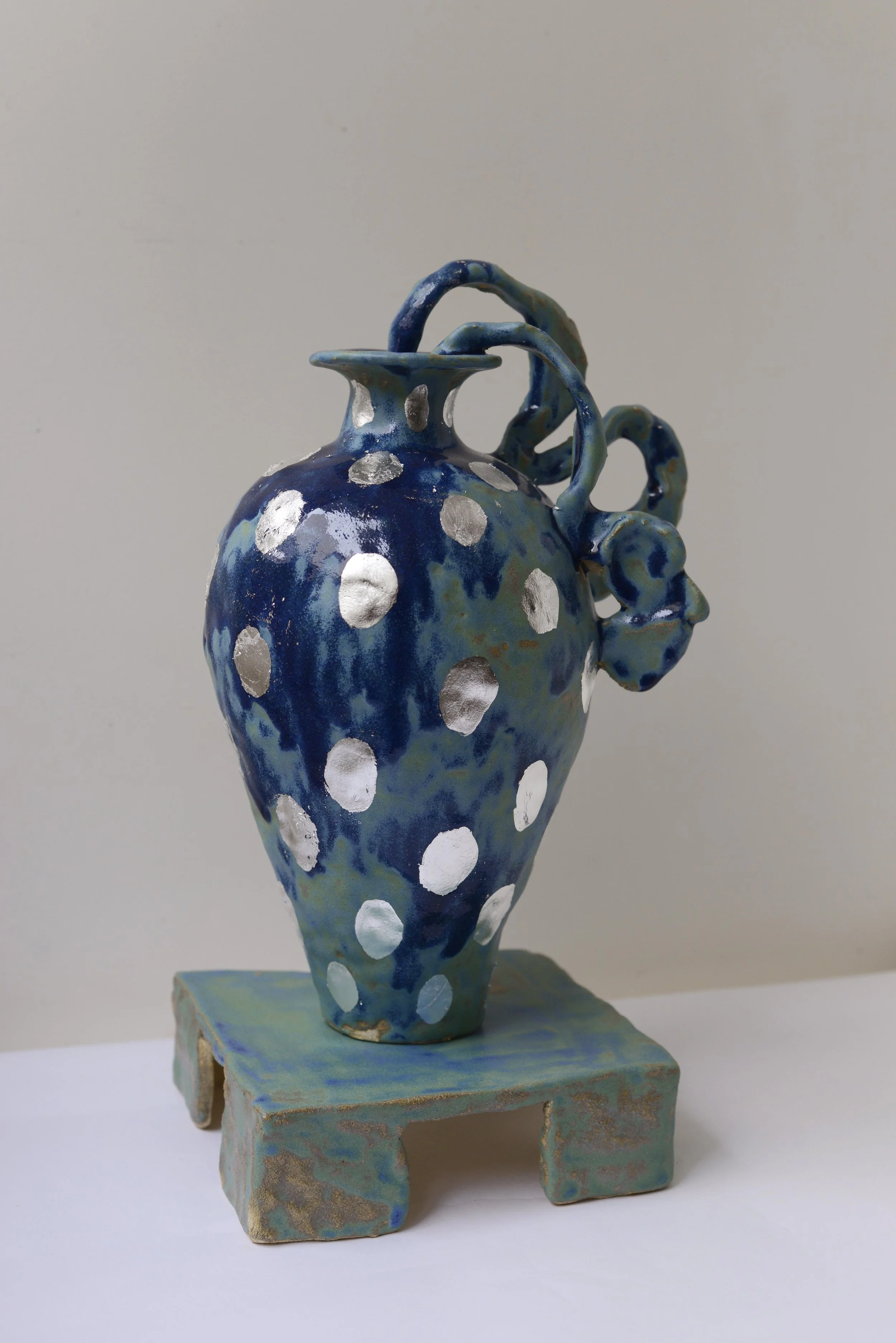 Une sculpture en céramique représentant un vase bleu avec des points métalliques argentés, posé sur une base en pierre. La sculpture a des formes ondulantes et des caractéristiques artistiques abstraites.