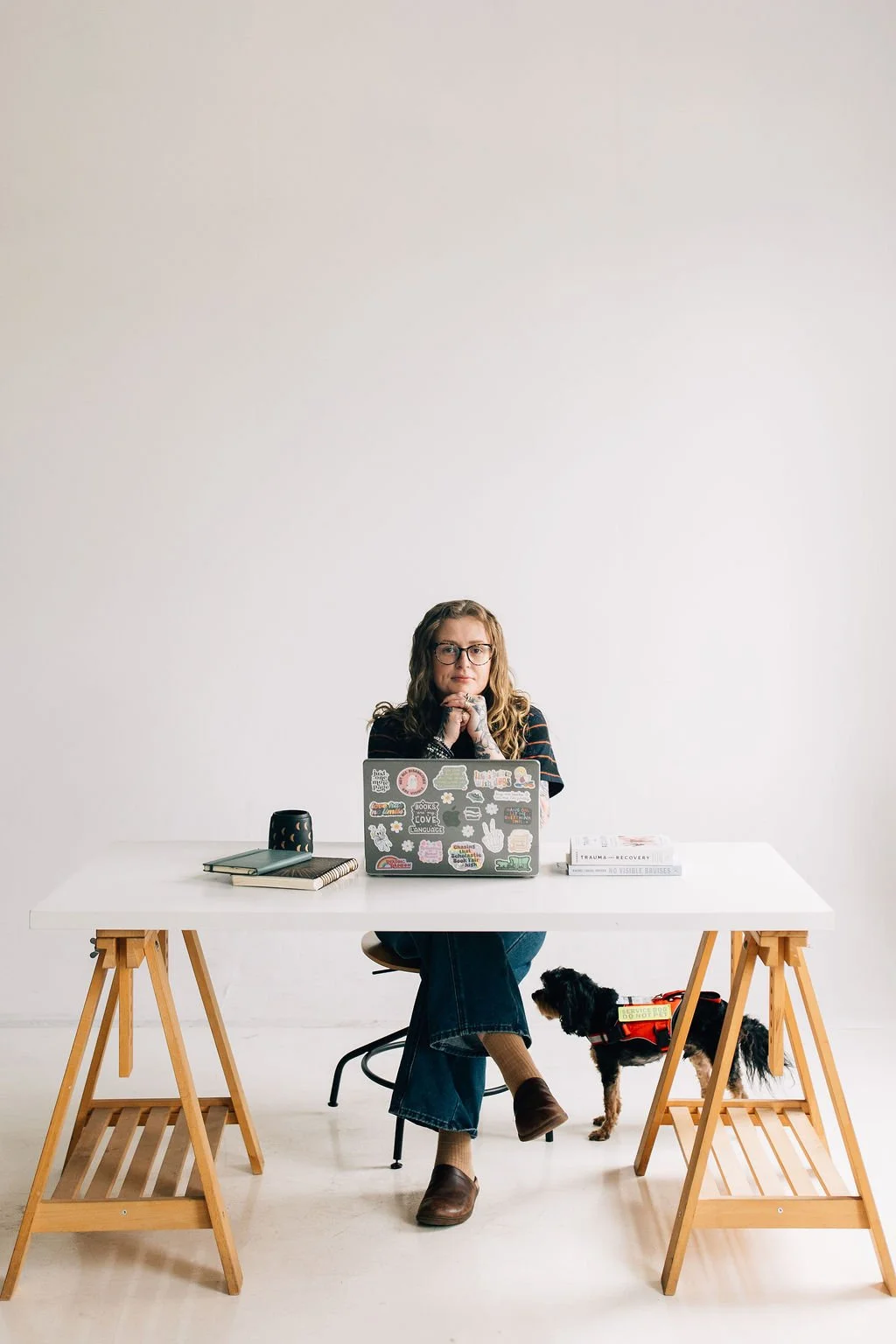 A woman sitting at a white desk with a laptop, notebooks, and a cup, with a small black and tan dog beside her. The woman is wearing glasses, a dark striped shirt, jeans, and brown boots, and is looking at the camera with her hands folded under her chin.