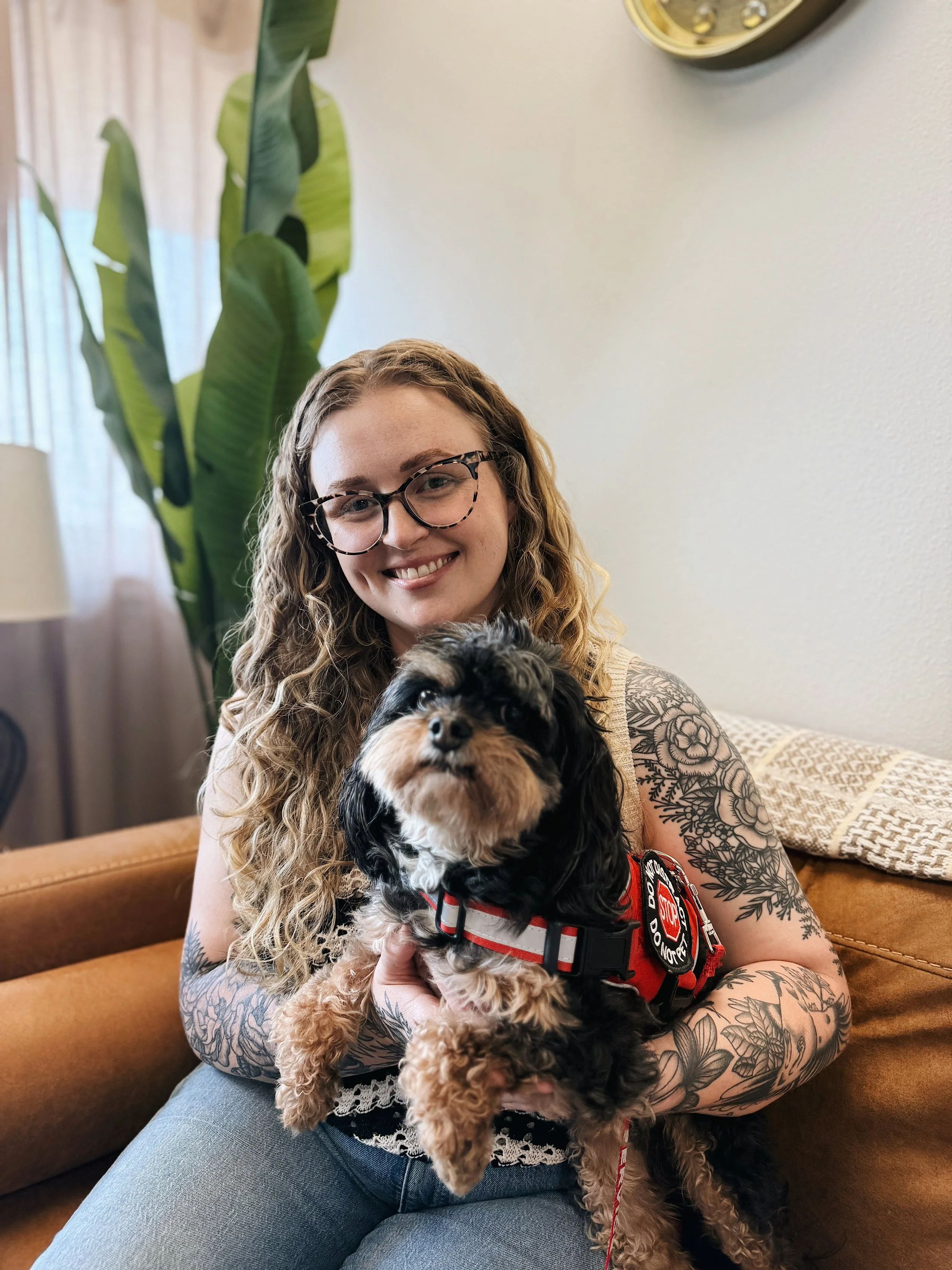 A smiling woman with long curly hair, wearing glasses and tattoos on her arms, holding a black and brown dog with a red harness indoors with a large green plant and a yellow wall clock in the background.
