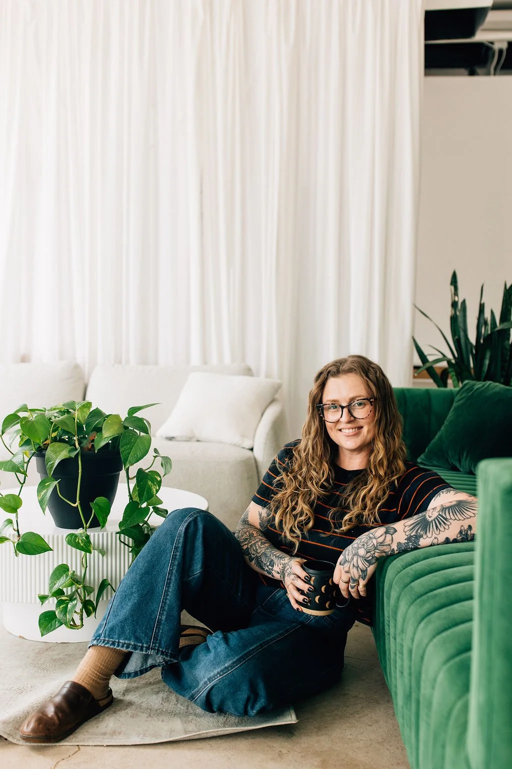 Colorado Trauma therapist Chaucee sitting on the ground with legs crossed holding a cup of coffee and leaning against a green jewel toned couch