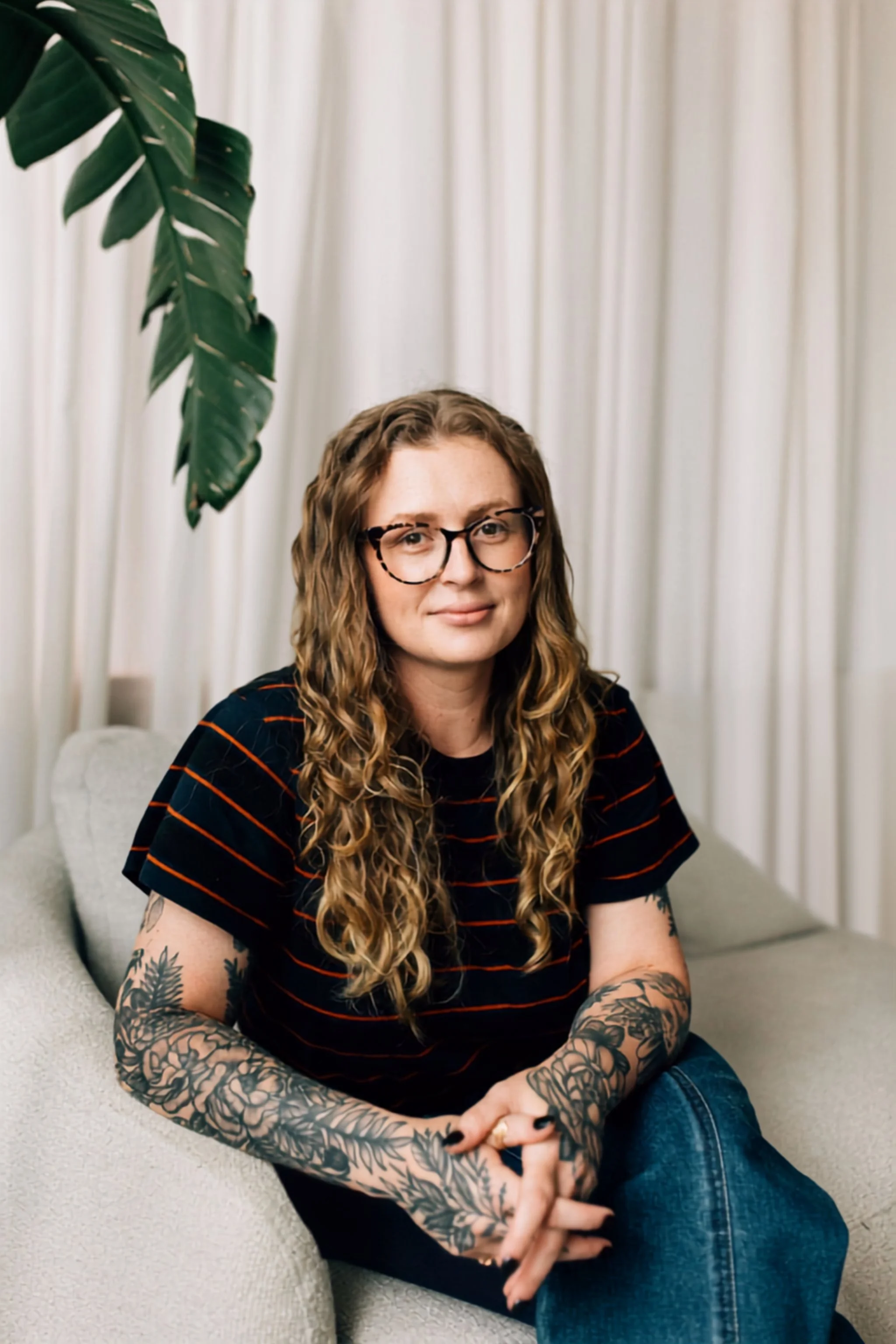 A woman with long curly hair and glasses sitting on a light-colored sofa, wearing a black T-shirt with orange stripes, and decorated with tattoos on her arms, against a backdrop of white curtains and a large green plant.