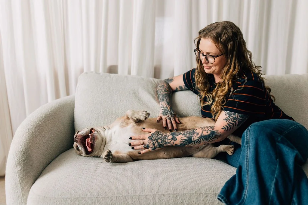 A woman with tattoos and glasses gently petting a yawning dog lying on a beige couch in a bright room with white curtains.