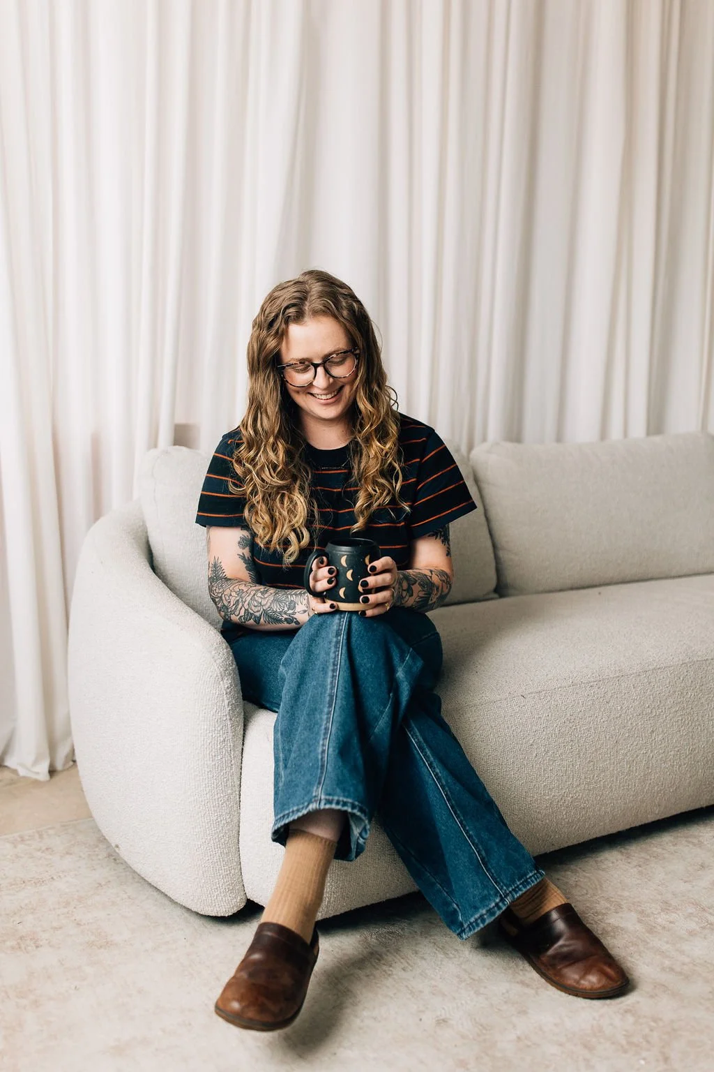 A young woman with glasses and tattoos smiling while sitting on a light-colored sofa, holding a black mug with a moon design, in a room with white curtains.