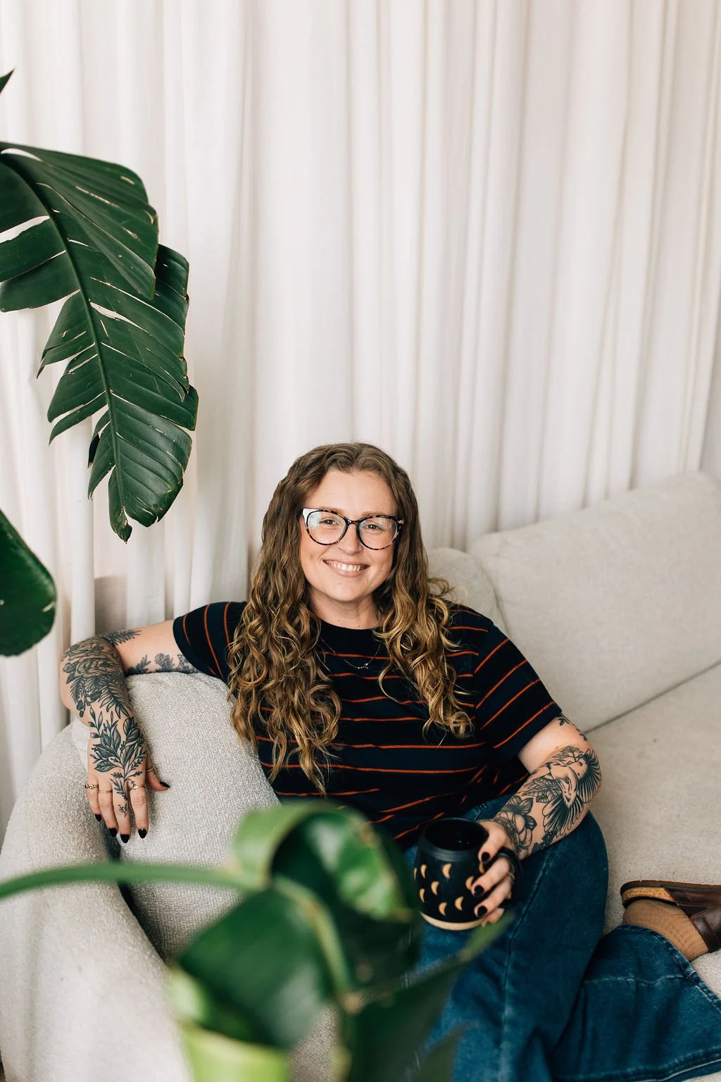 A woman with long curly hair, wearing glasses, a black t-shirt with red stripes, and tattoos on her arms, sitting on a cream couch, holding a black mug with a moon phase pattern, smiling at the camera.