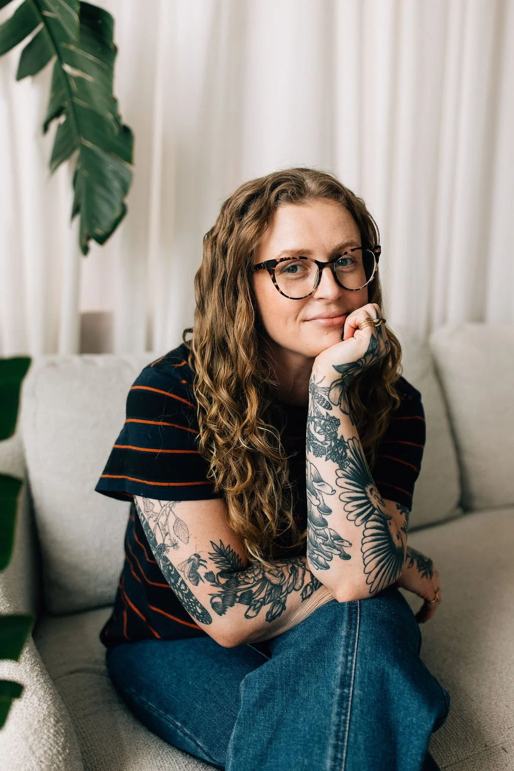 A woman with wavy, long hair, wearing large glasses and a striped black and orange shirt, sitting on a light-colored sofa, looking at the camera with a slight smile, resting her chin on her hand, with tattoos on her arms, in a cozy indoor setting.