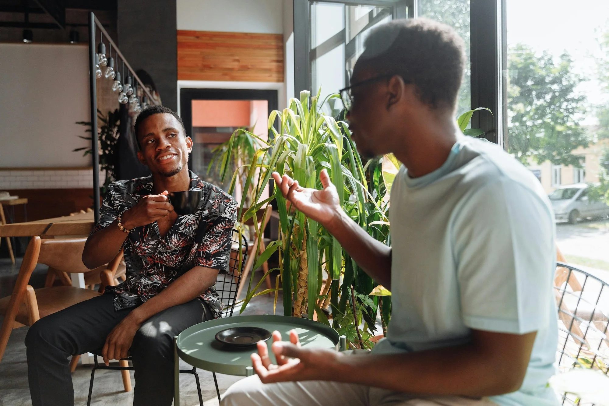Two young men are sitting and talking inside a cafe, one holding a black mug, the other gesturing with his hands. There are large windows with sunlight and greenery outside, and a potted plant between them.