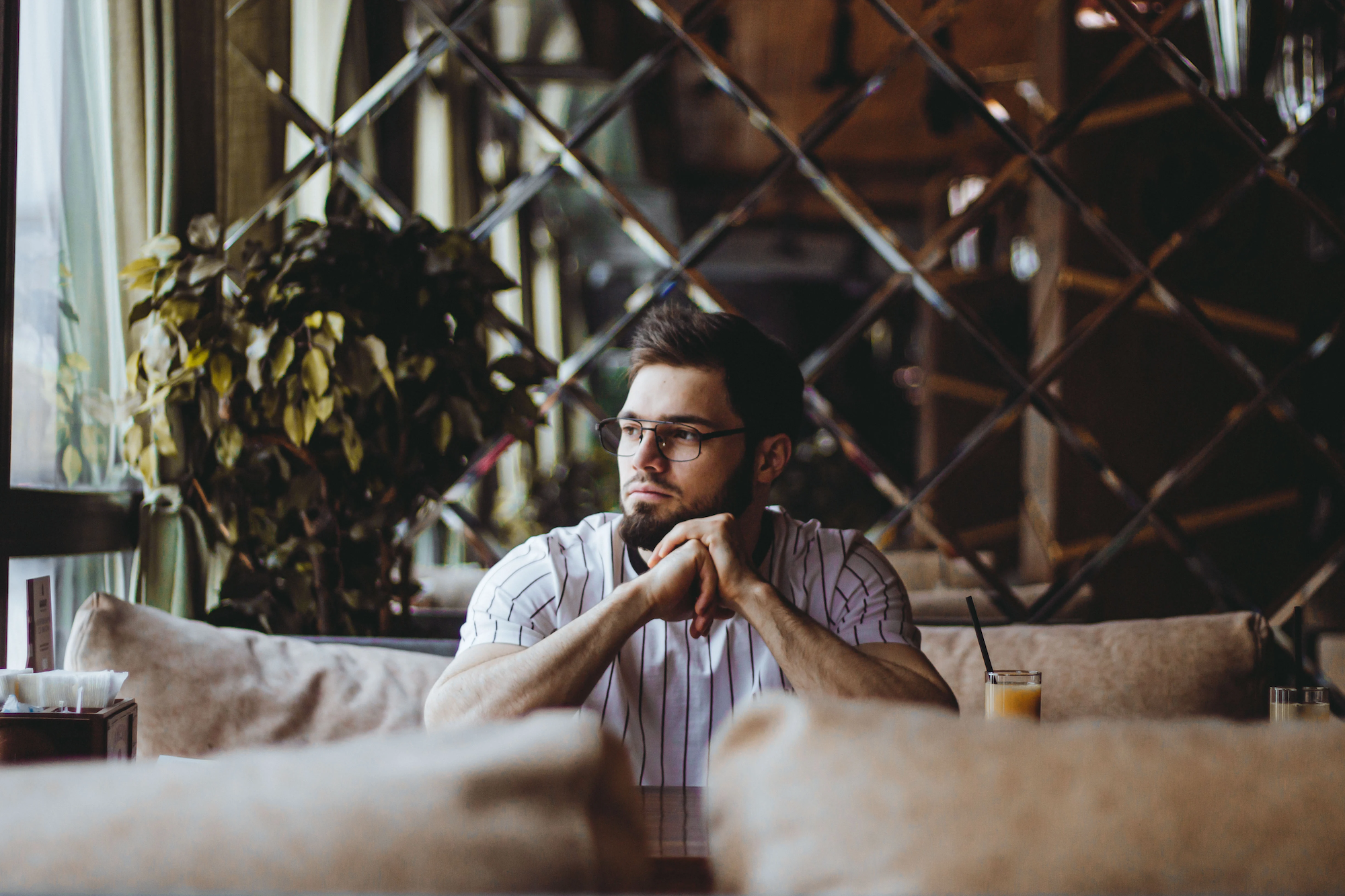 A man sitting at a table in a cozy, dimly lit cafe with a wooden interior and large windows, resting his chin on his hands and looking contemplative.