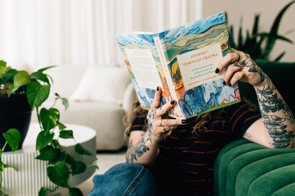 Chaucee, a trauma therapist in Colorado, is sitting on the floor, reading a book on trauma, holding it up in front of her face while leaning against a green velvet couch