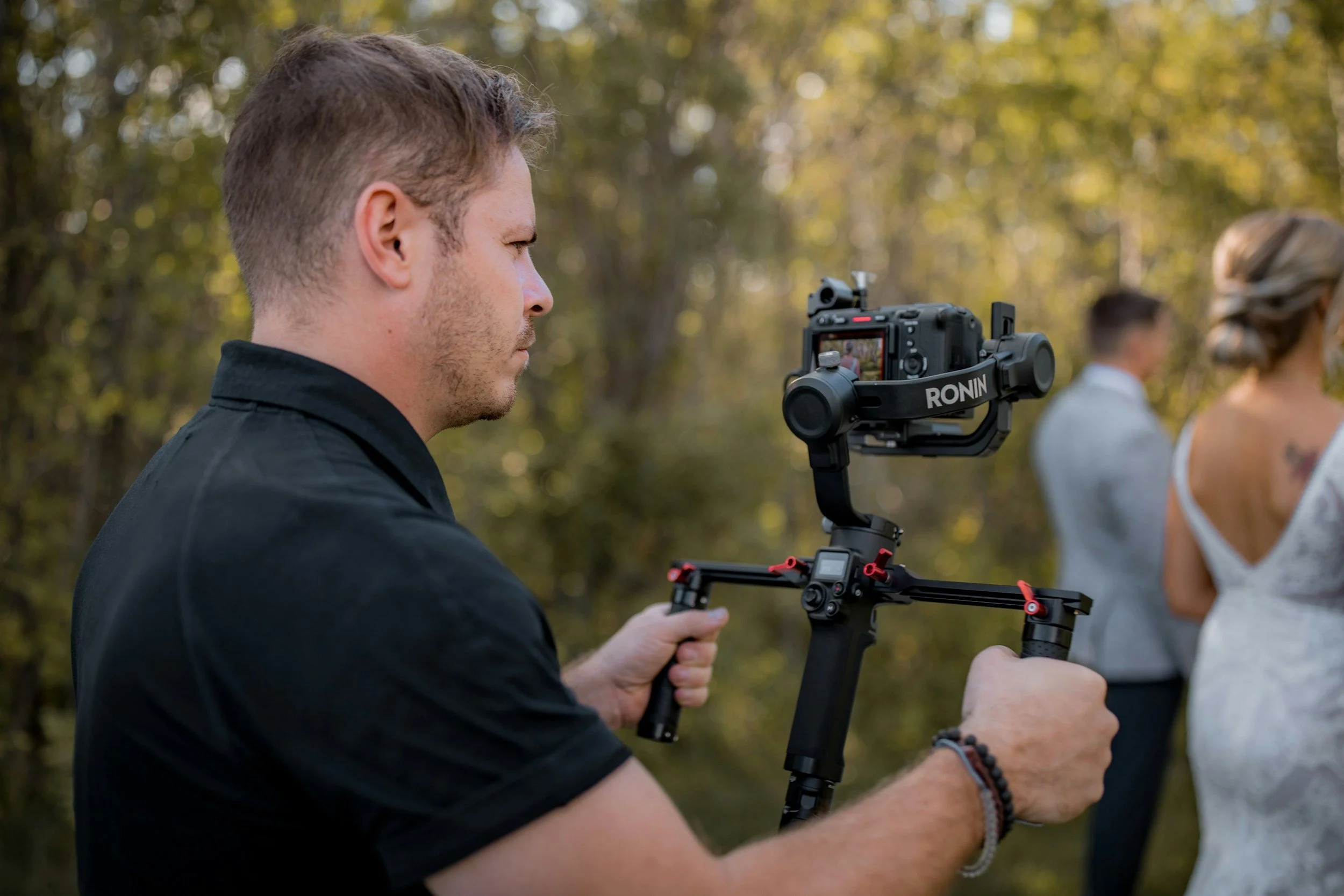 A man using a camera stabilizer to record a scene outdoors during daytime, with two women in the background, one with blonde hair in a bun and the other with dark hair, in a wooded area.