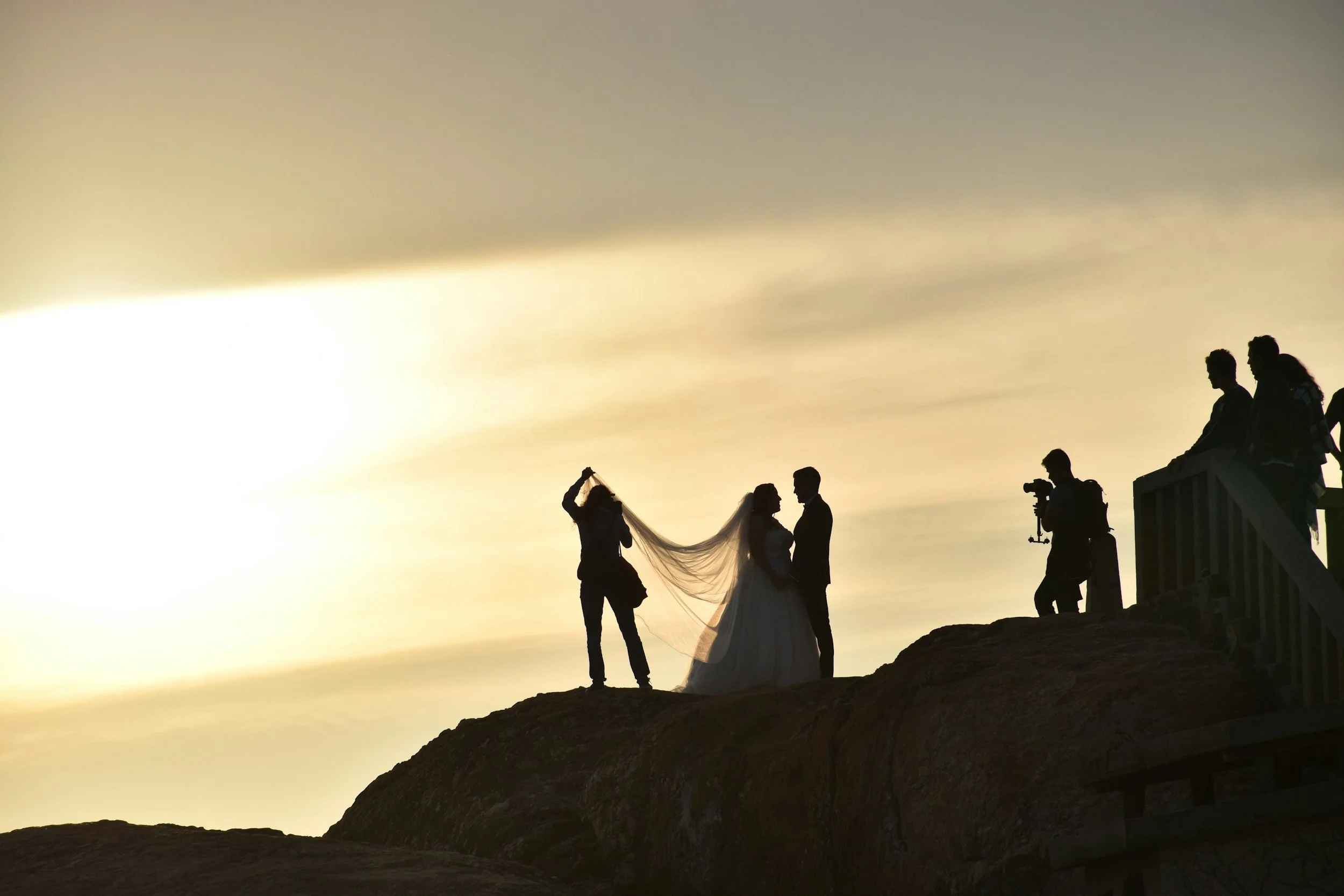 Silhouette of a bride and groom standing on a hill at sunset, with a woman holding a veil and a photographer with a camera on a tripod nearby.