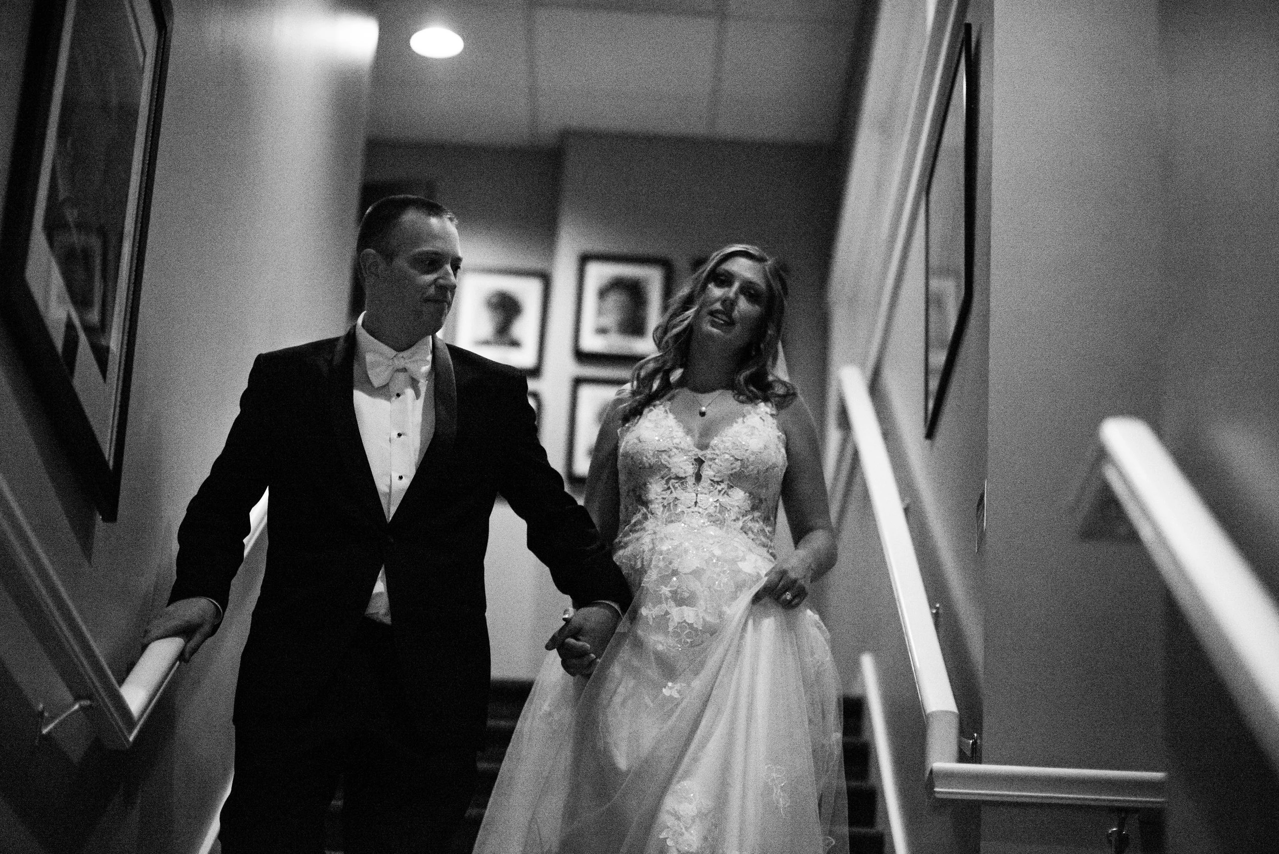 A black and white photo of a bride and groom walking down a staircase, holding hands. The groom is wearing a tuxedo and the bride is wearing a lace wedding dress. They are inside a building with framed artwork on the walls.