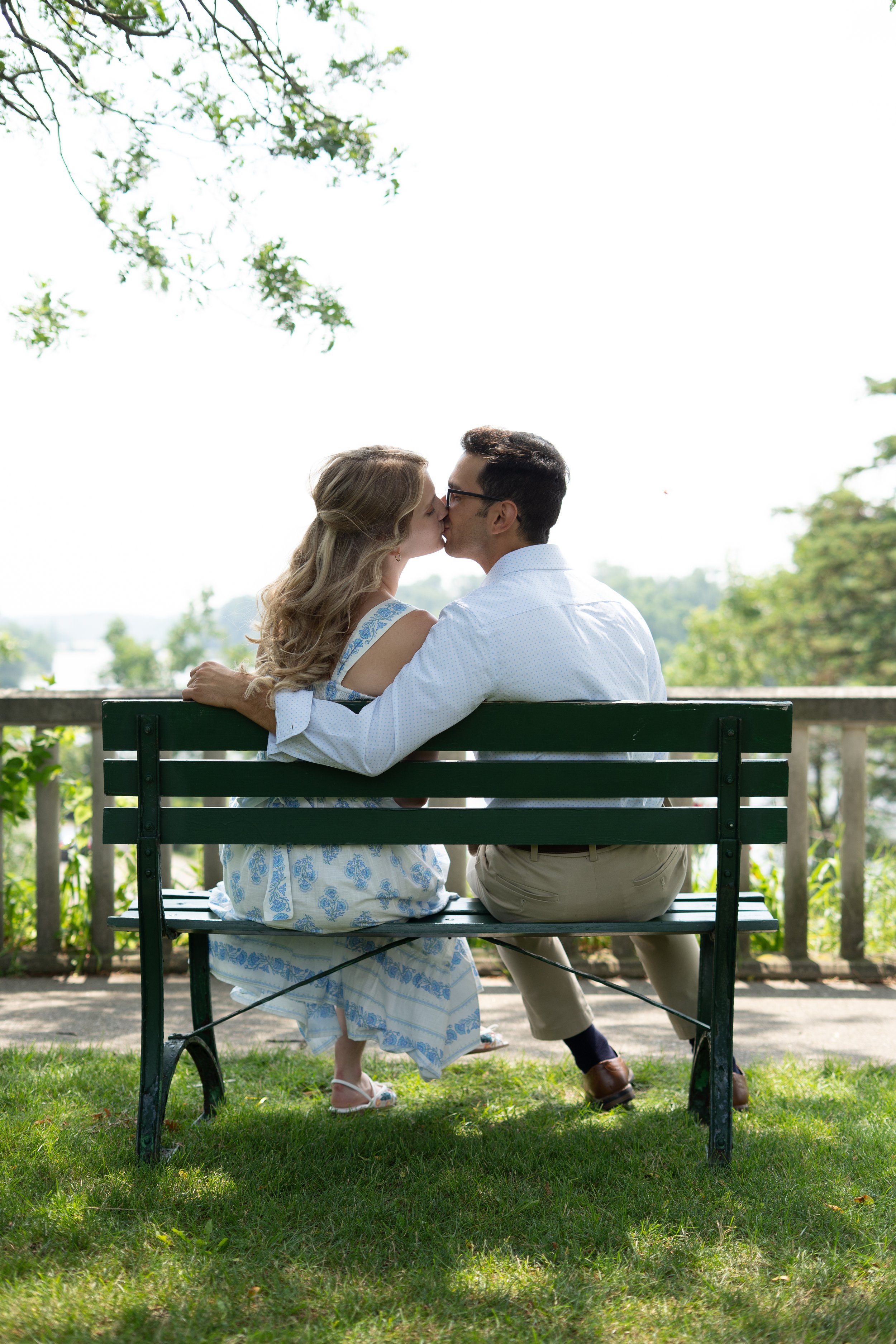 A couple sharing a kiss on a park bench, surrounded by trees and greenery, with the sky visible in the background.