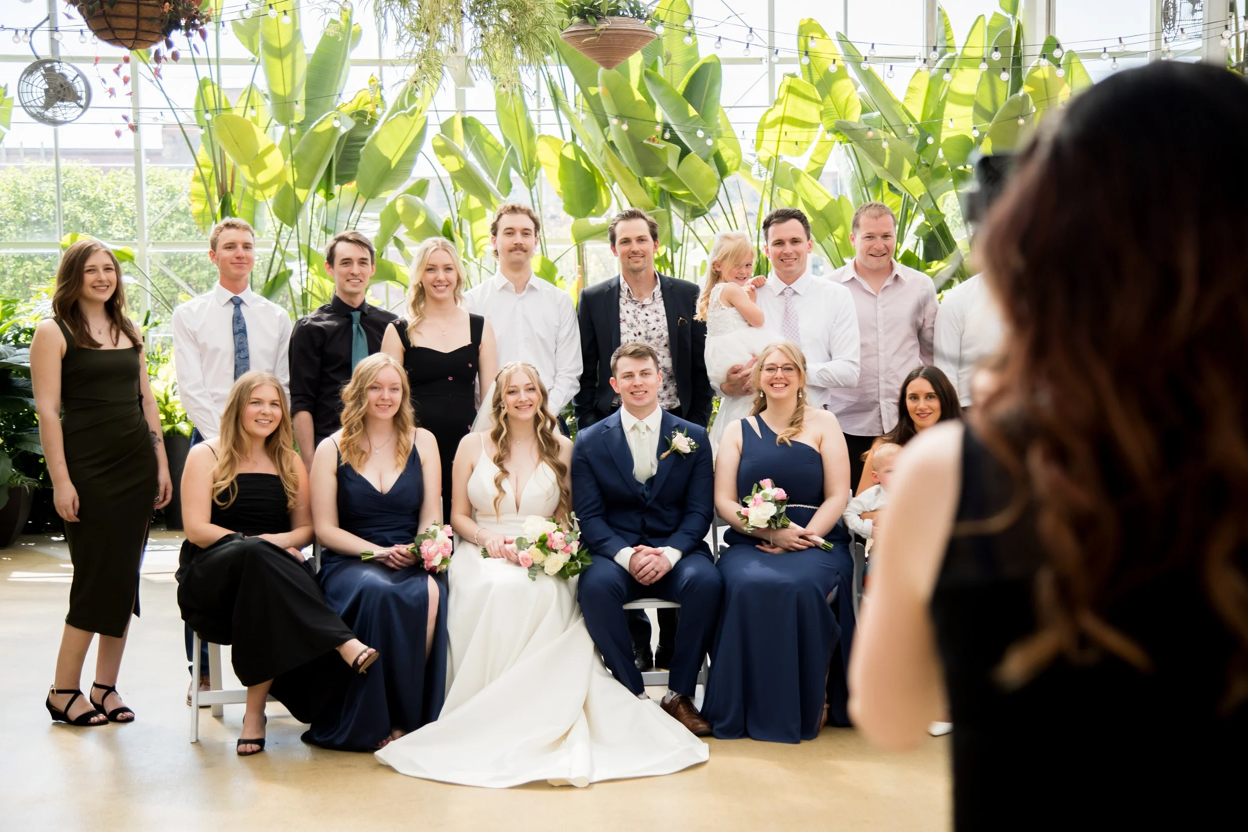 A group of people at a wedding ceremony, with the bride and groom seated in front and surrounded by wedding party members, in a bright greenhouse with large green leaves and string lights overhead.