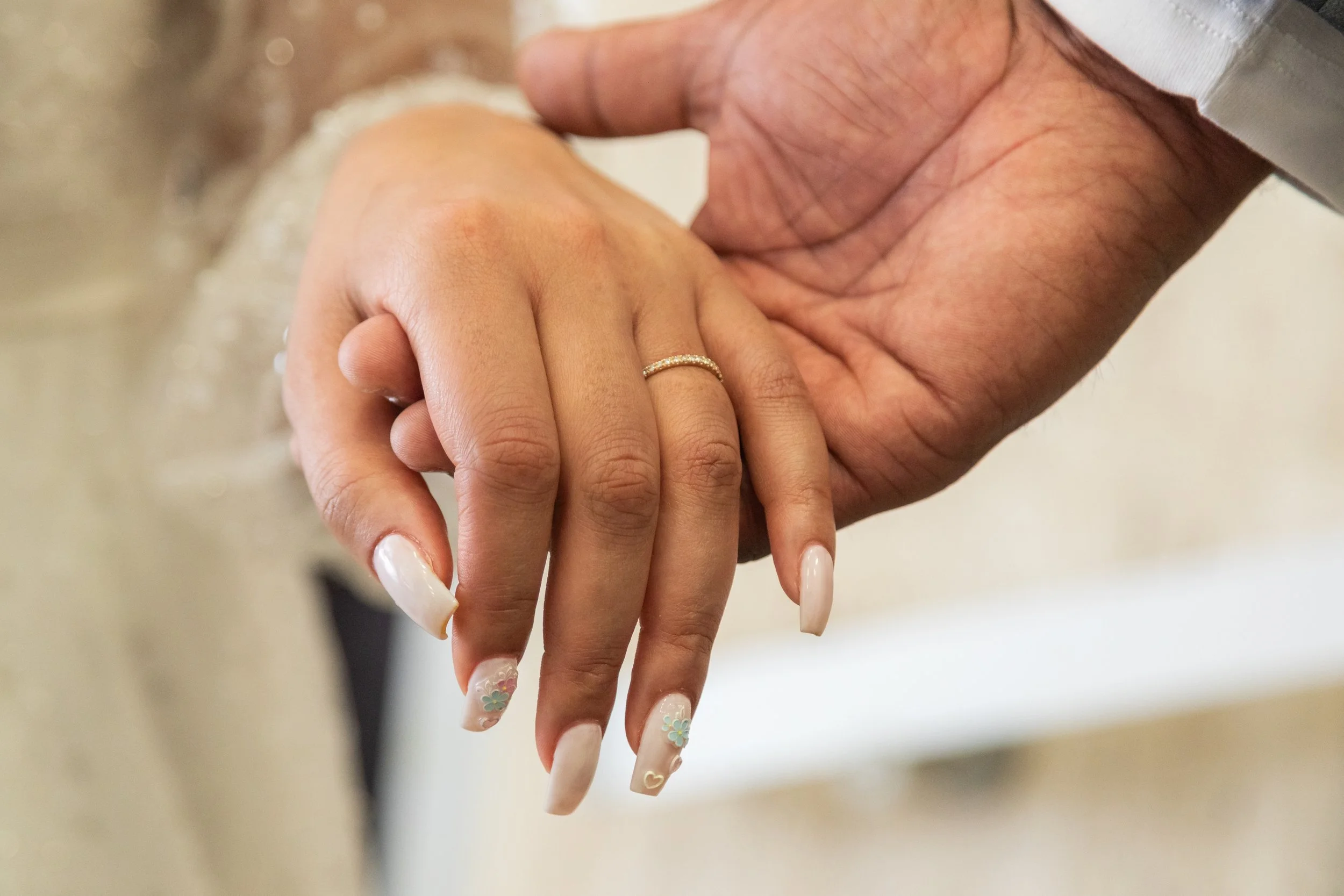 Close-up of a newlywed couple holding hands, showing the bride's manicured nails with flower designs and a wedding ring on her finger, and the groom's hand with visible wrinkles and a white shirt cuff.